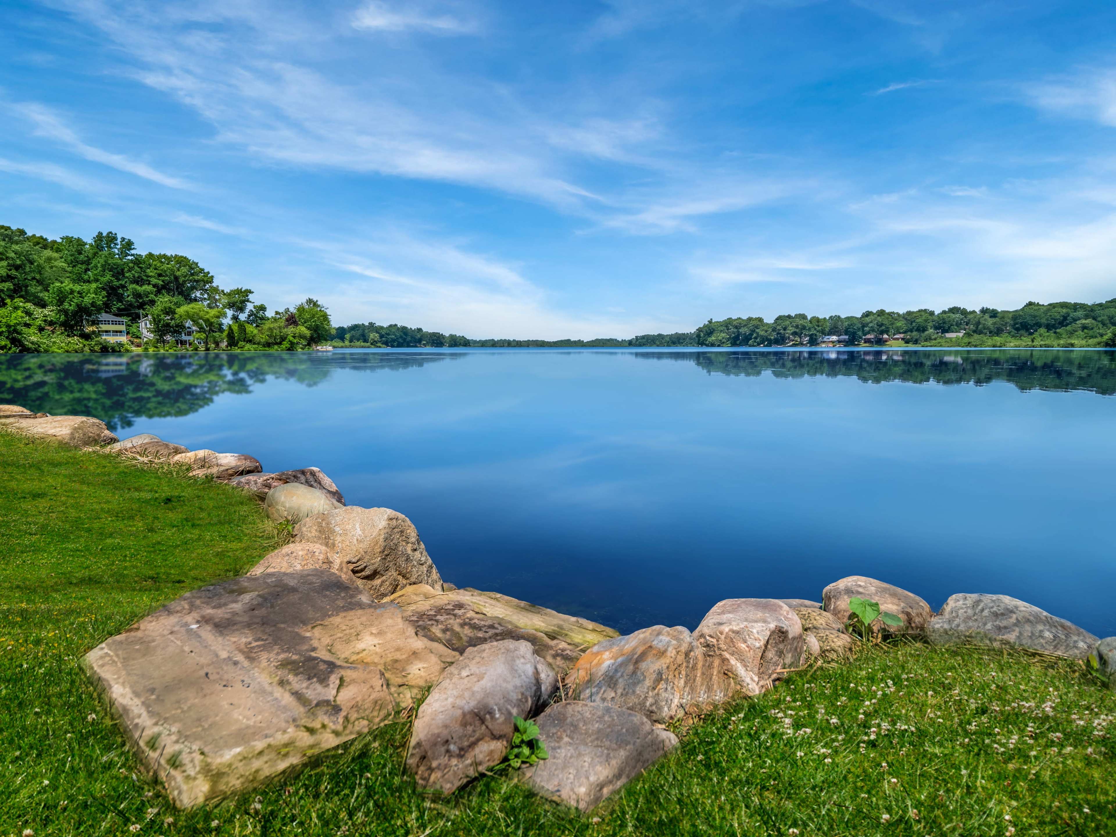 A calm lake reflects the clear blue sky and surrounding greenery, bordered by a rocky edge and grassy area.