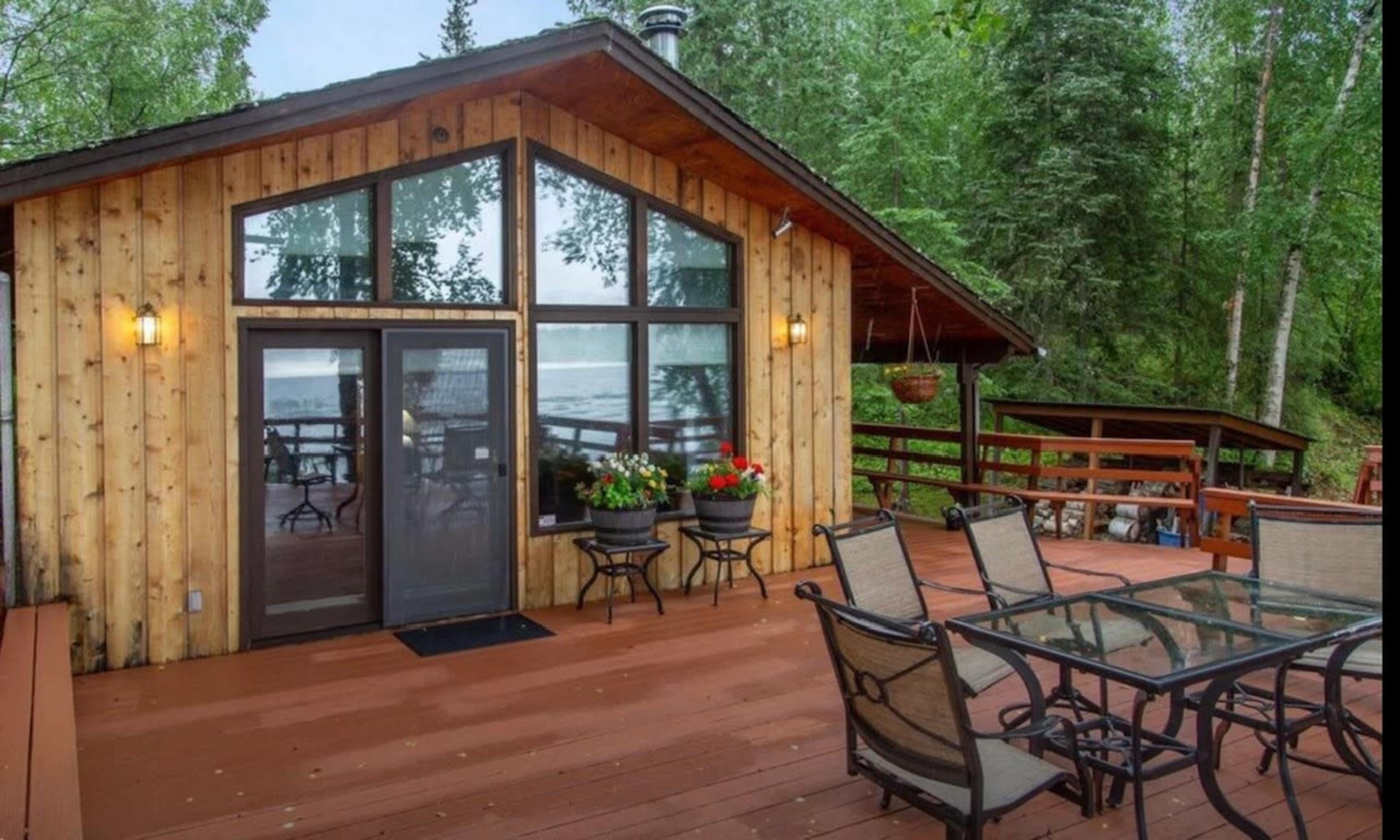 A wooden cabin with large windows overlooks a deck furnished with a glass table and chairs, surrounded by trees near a body of water.