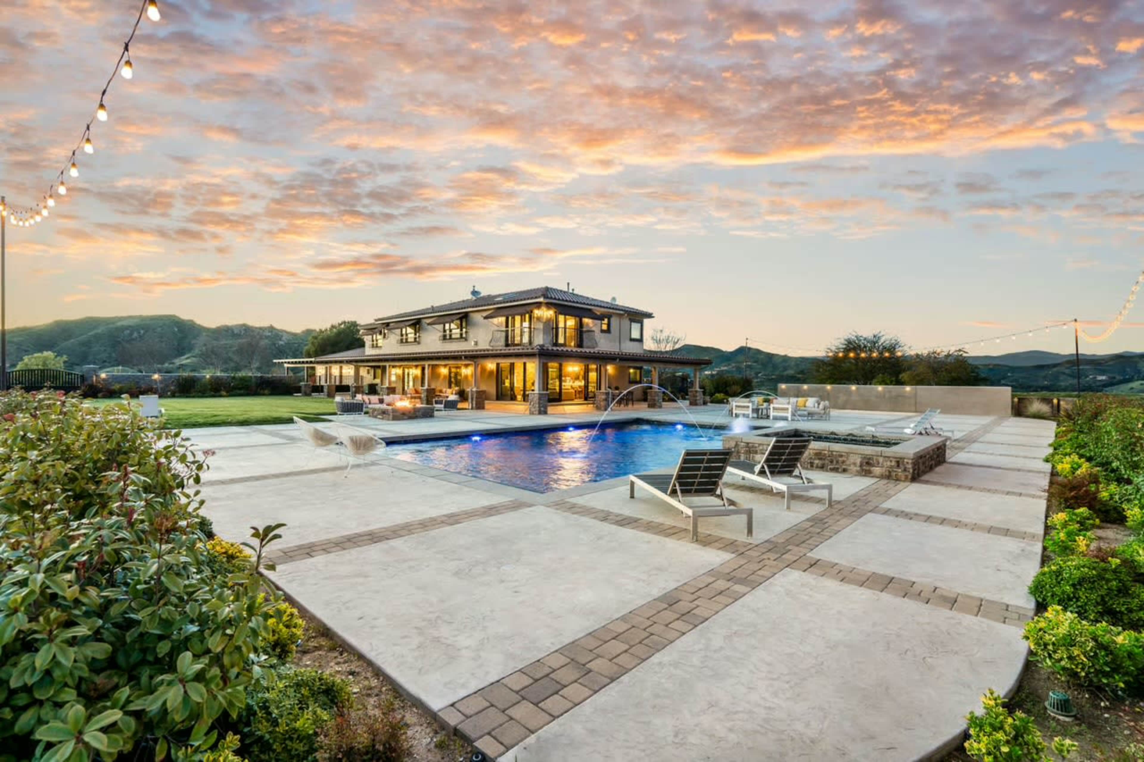 A modern two-story house with large windows overlooks a swimming pool surrounded by a stone patio, set against a backdrop of a colorful sunset and rolling hills.