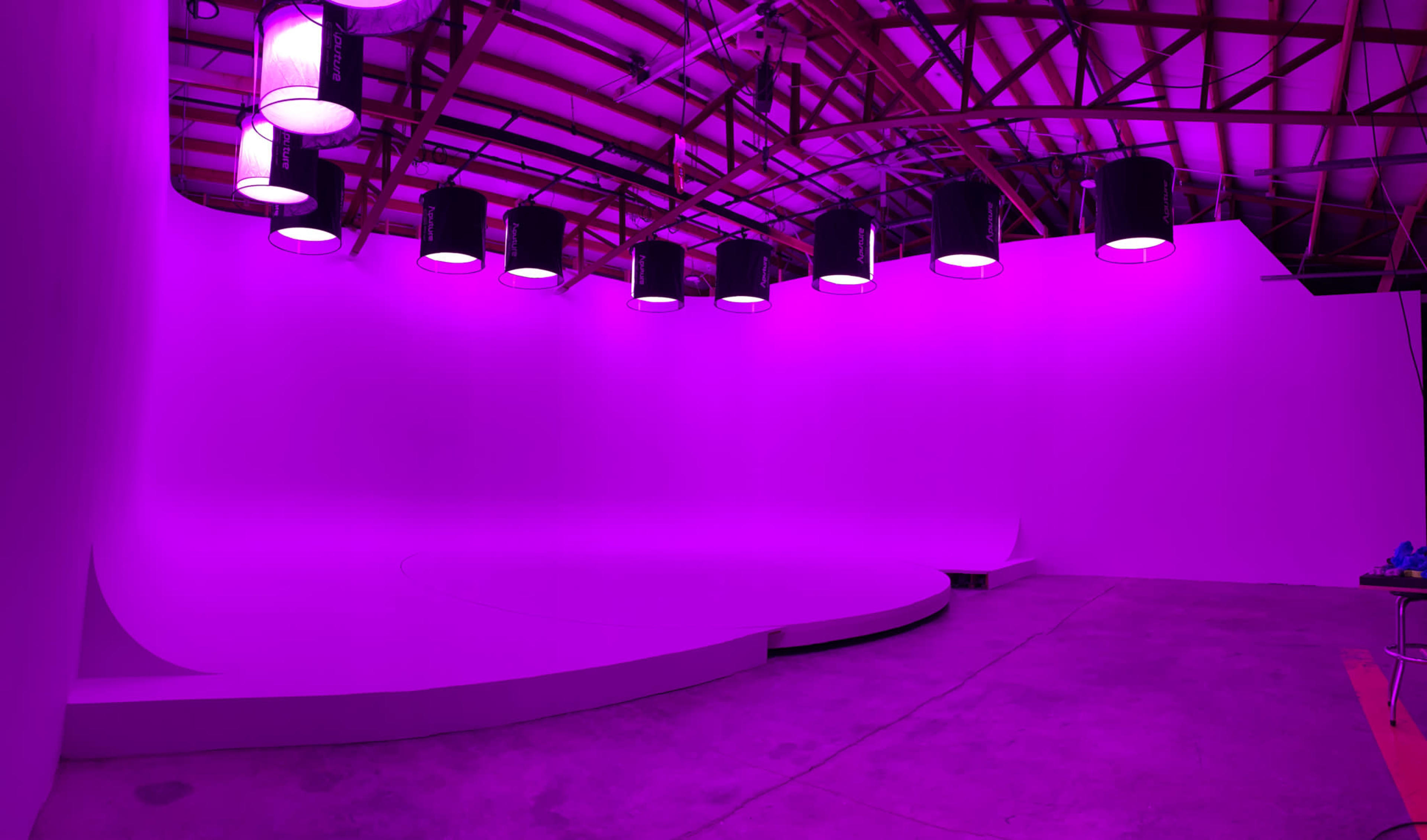The image shows a spacious studio with a curved platform and multiple pendant lights mounted on the ceiling, all illuminated in purple light.
