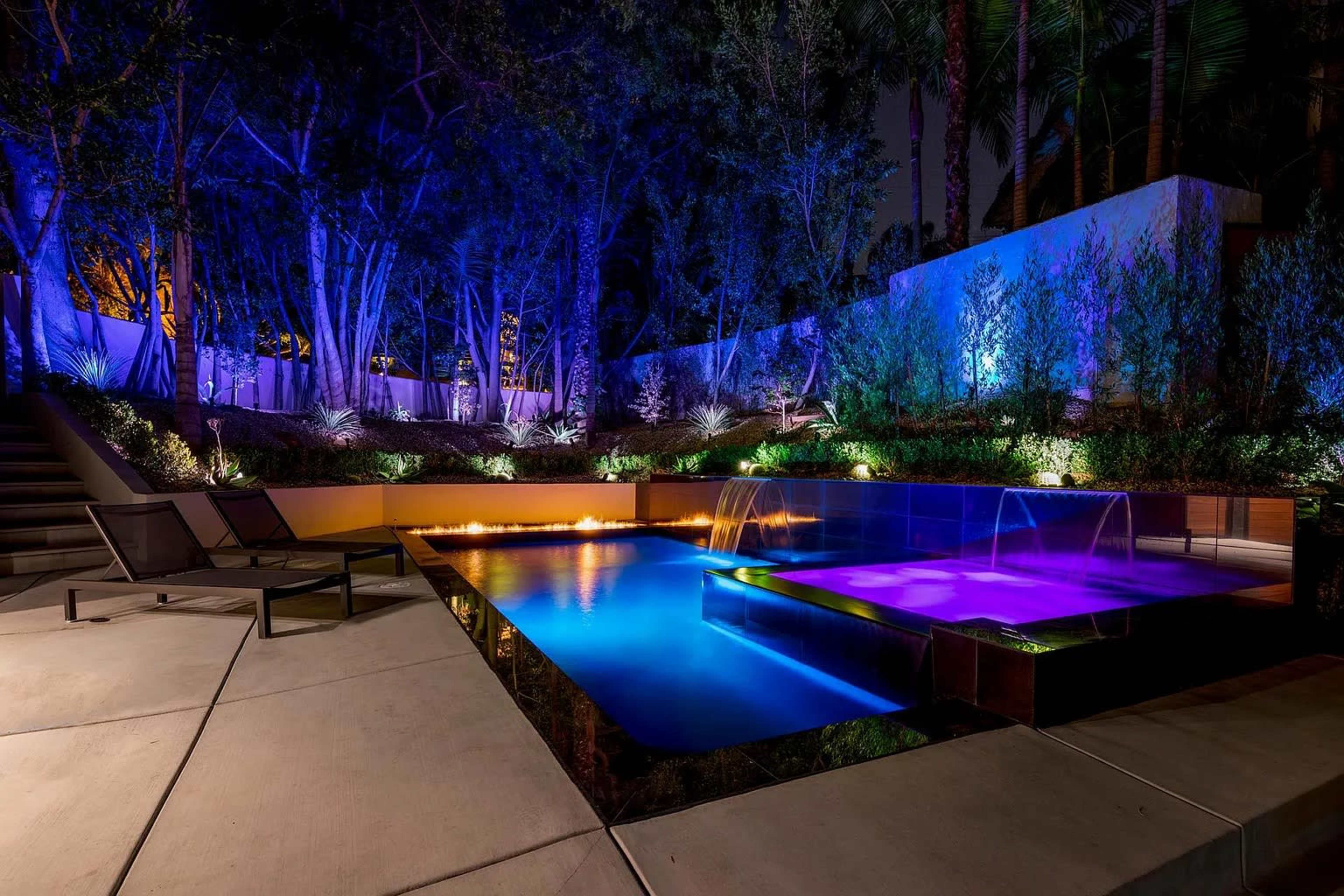 The image shows a modern swimming pool illuminated with blue and purple lights, surrounded by lush greenery and a concrete patio with loungers.