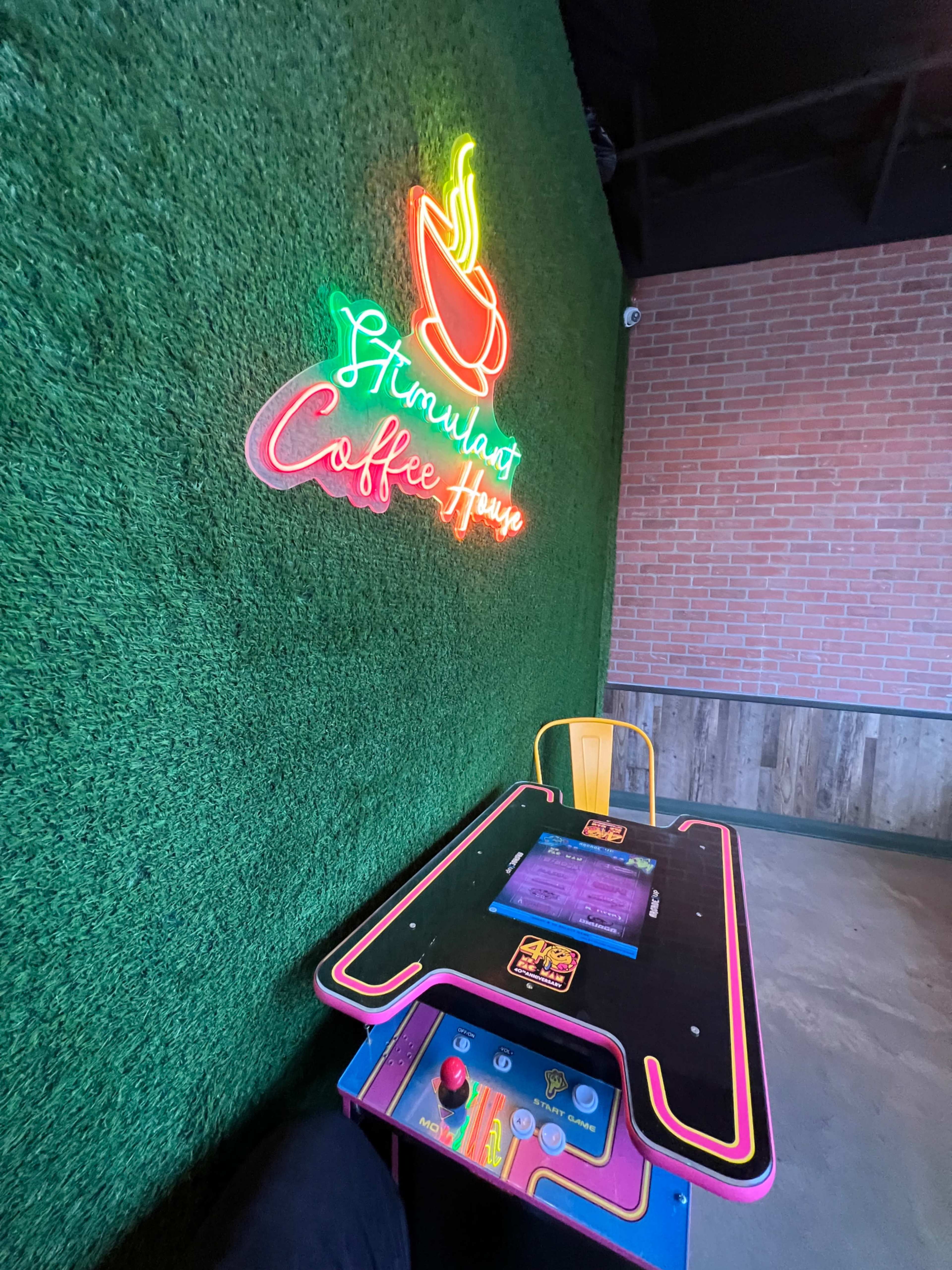 A brightly lit coffee house features a neon sign against a green wall and a retro arcade-style table.