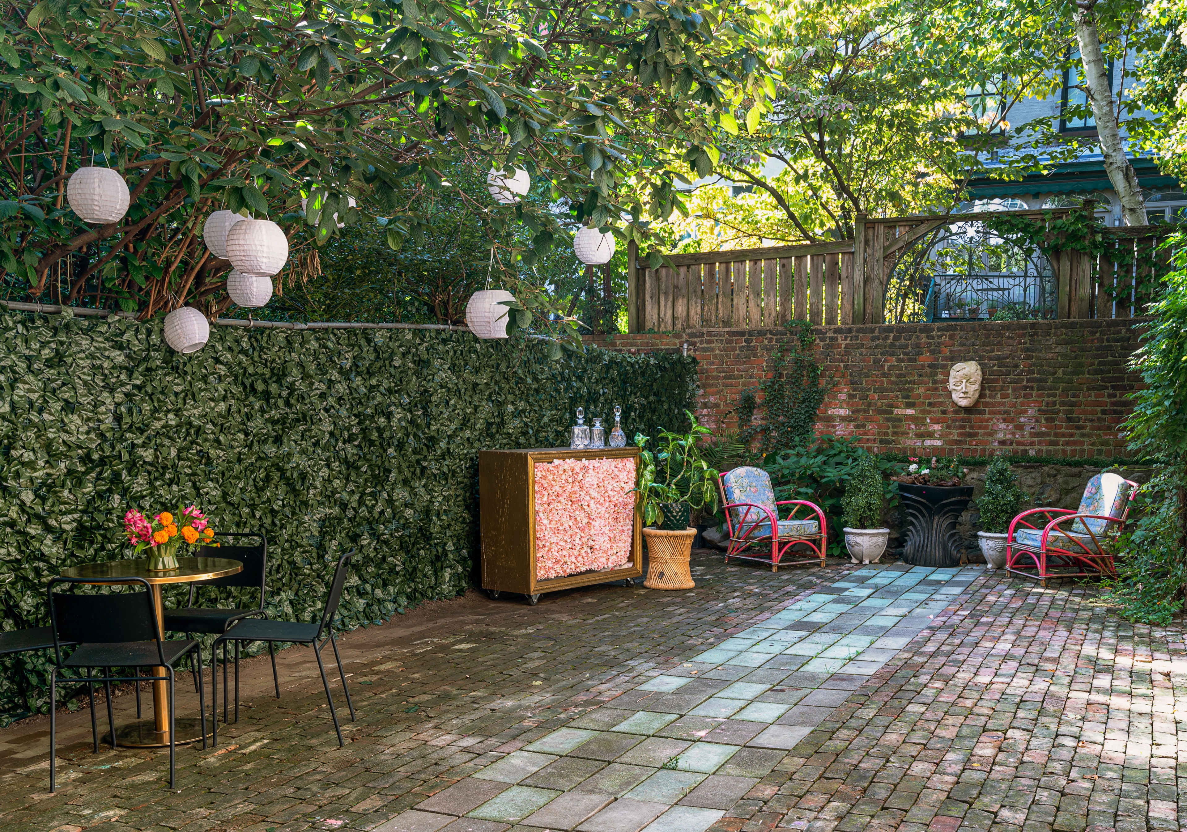 A shaded backyard garden features a stone patio with a decorative wooden cabinet, outdoor seating, and hanging lanterns amidst lush greenery.