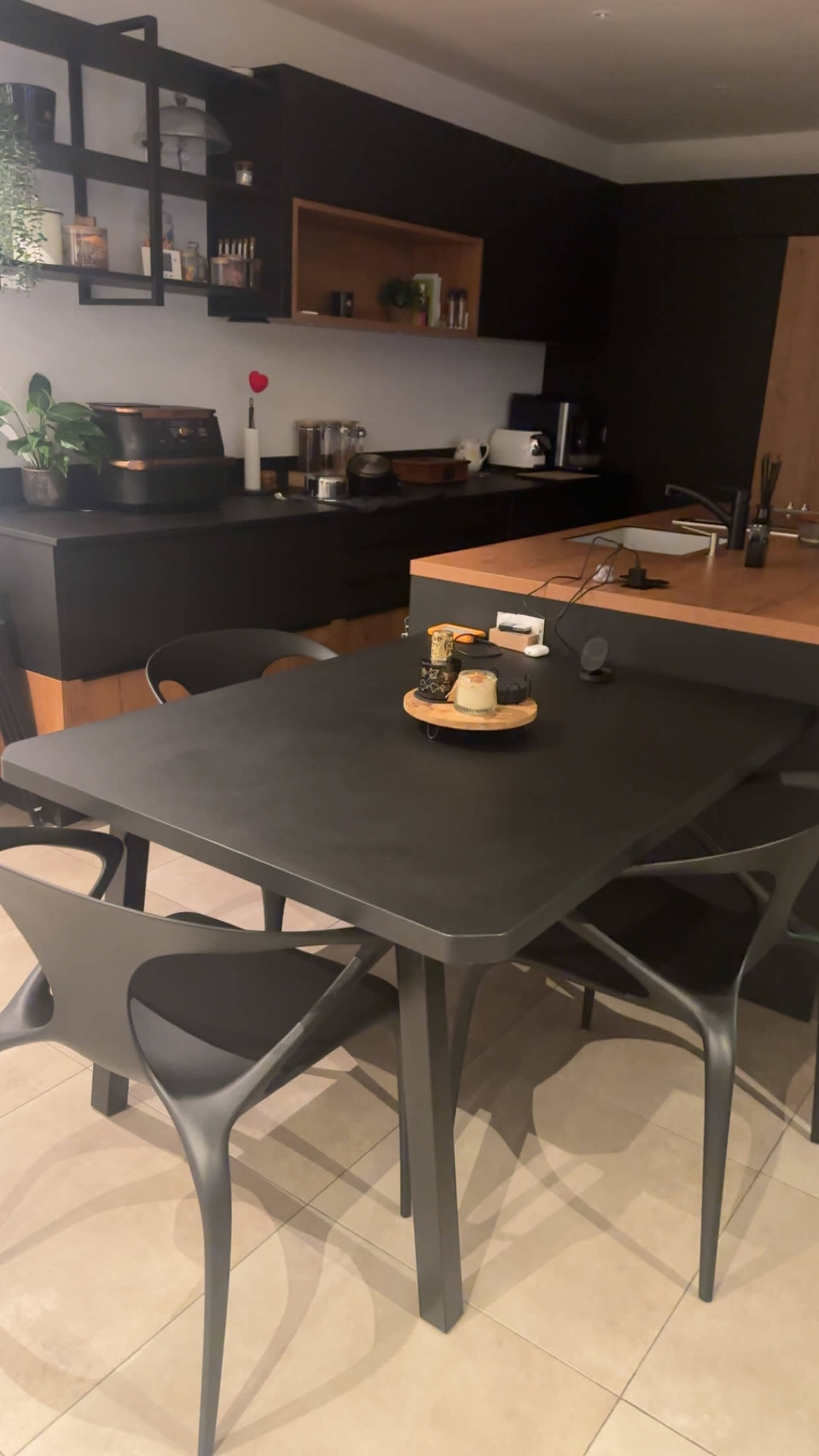 A modern kitchen features a dark table surrounded by black chairs, with a centerpiece and various kitchen appliances visible in the background.