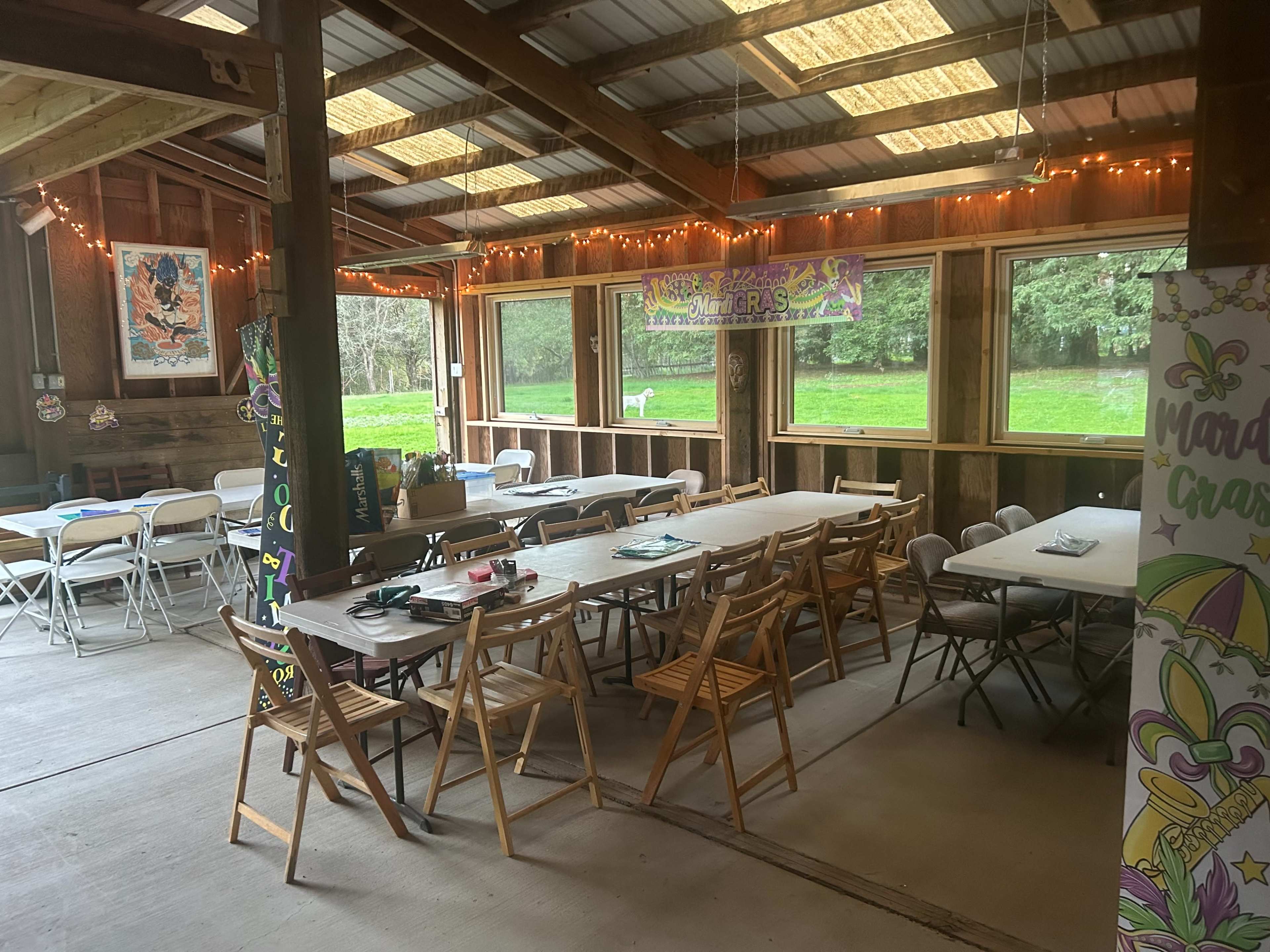 The image shows a spacious indoor gathering area with several tables and chairs arranged, decorated with colorful banners and string lights, and large windows offering a view of greenery outside.