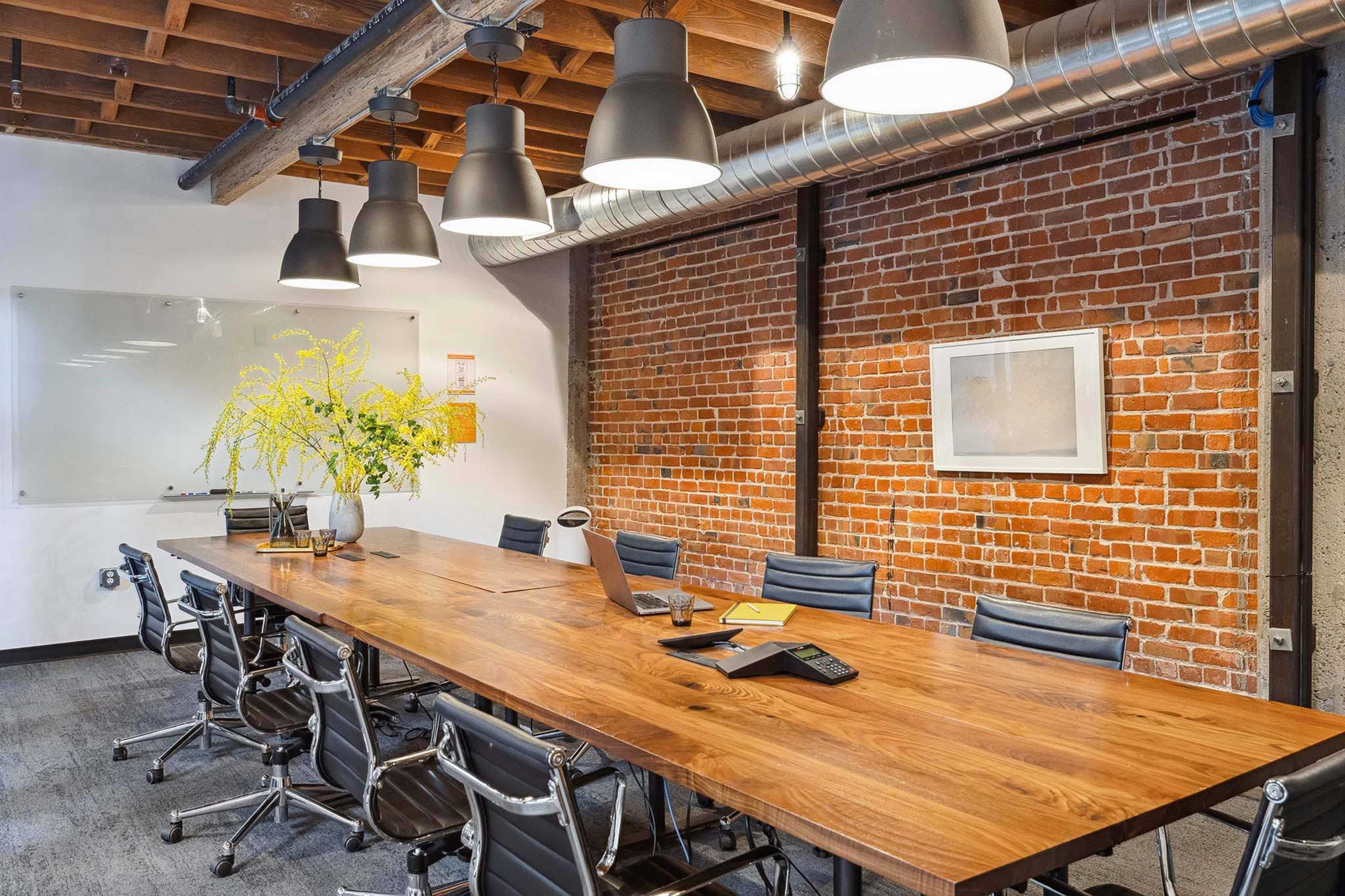 A large wooden conference table is surrounded by black office chairs in a well-lit meeting room with exposed brick walls and industrial lighting.