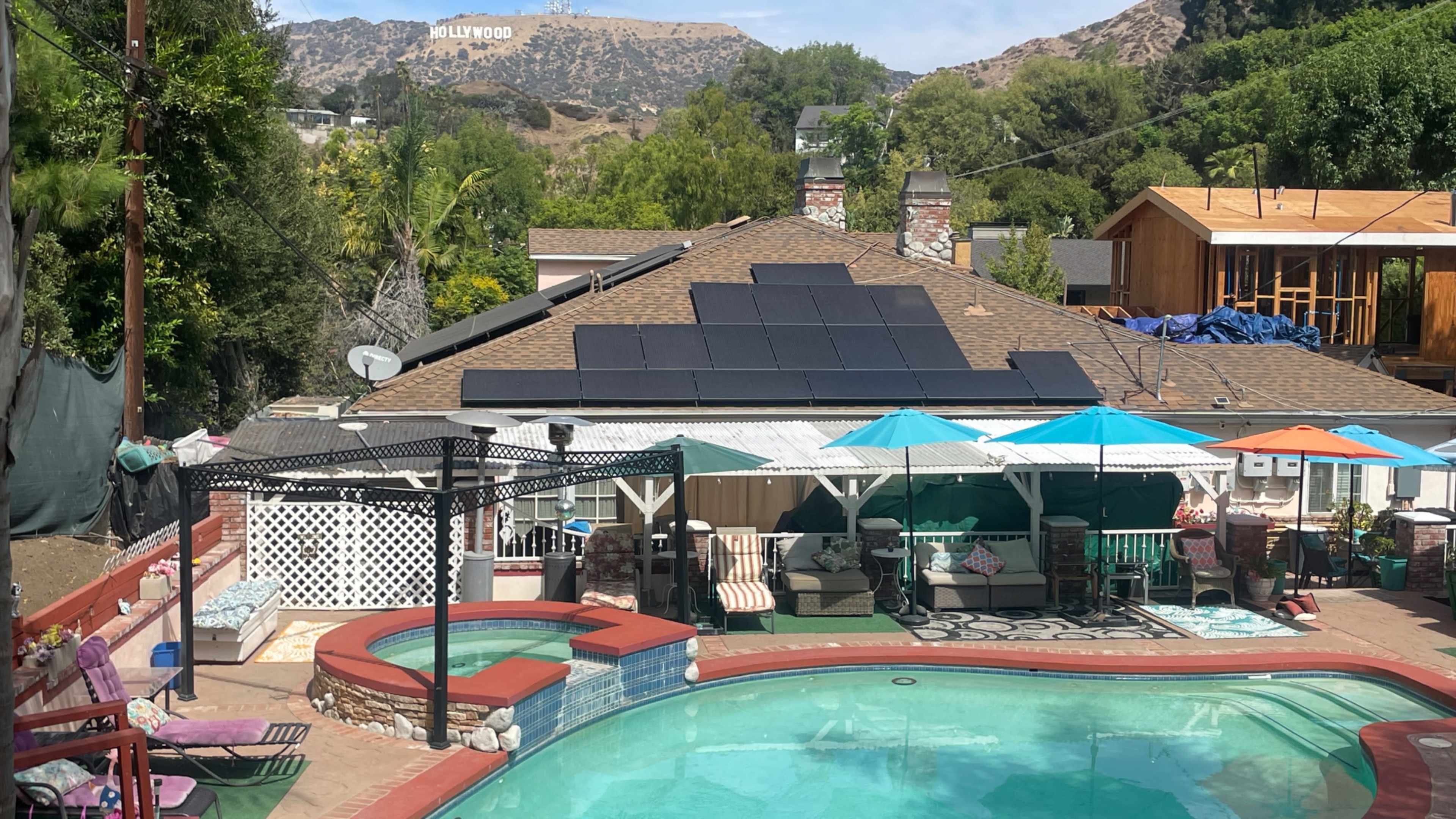 The image shows a residential backyard with a swimming pool, outdoor seating areas, and solar panels on the roof, with the Hollywood sign visible in the background.