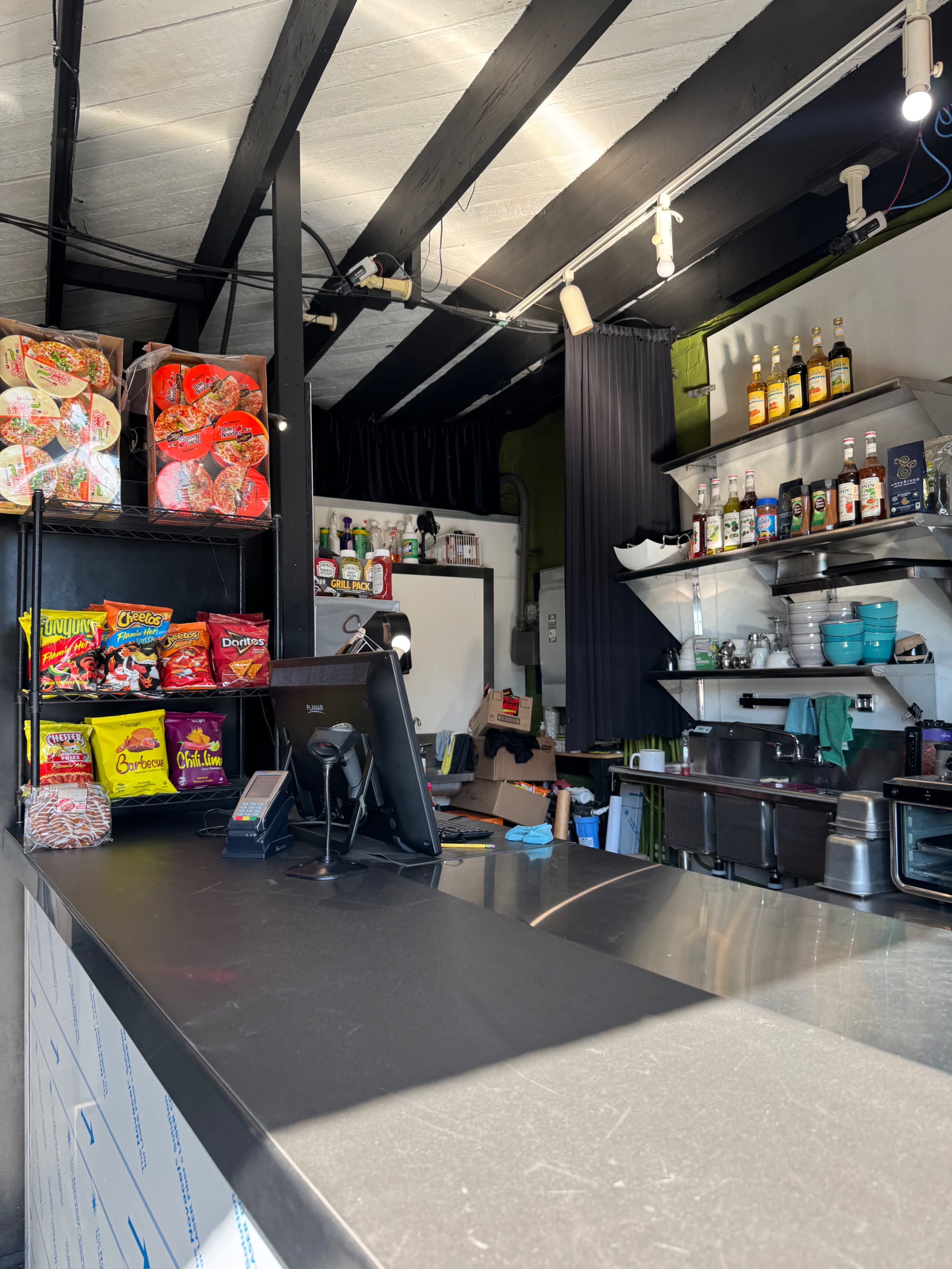 The image shows a small counter area of a store or café with snacks displayed on shelves and a cash register on the countertop.