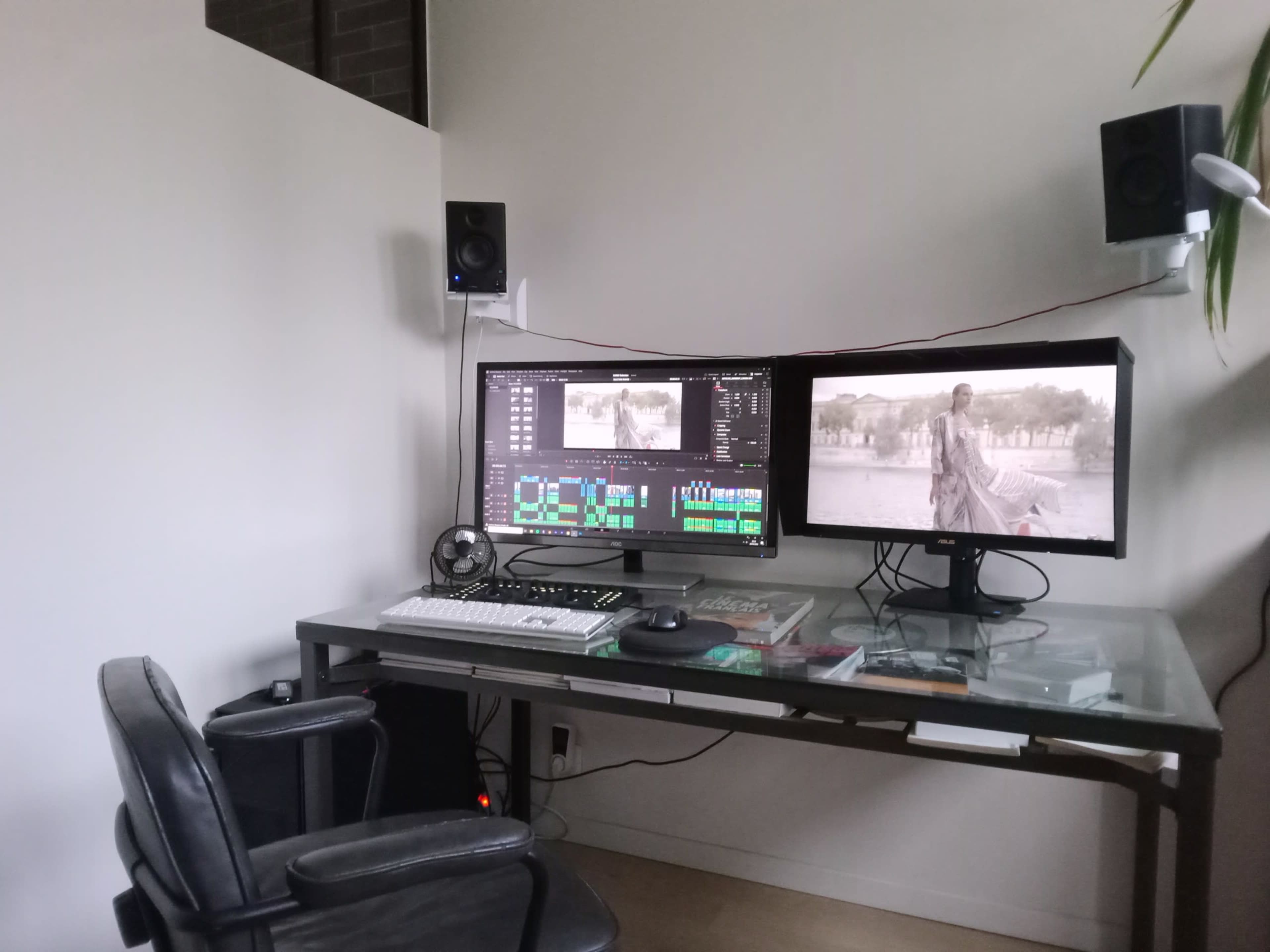 The image depicts a workspace featuring a glass desk with dual monitors, a keyboard, and a swivel chair, alongside speakers mounted on the wall.