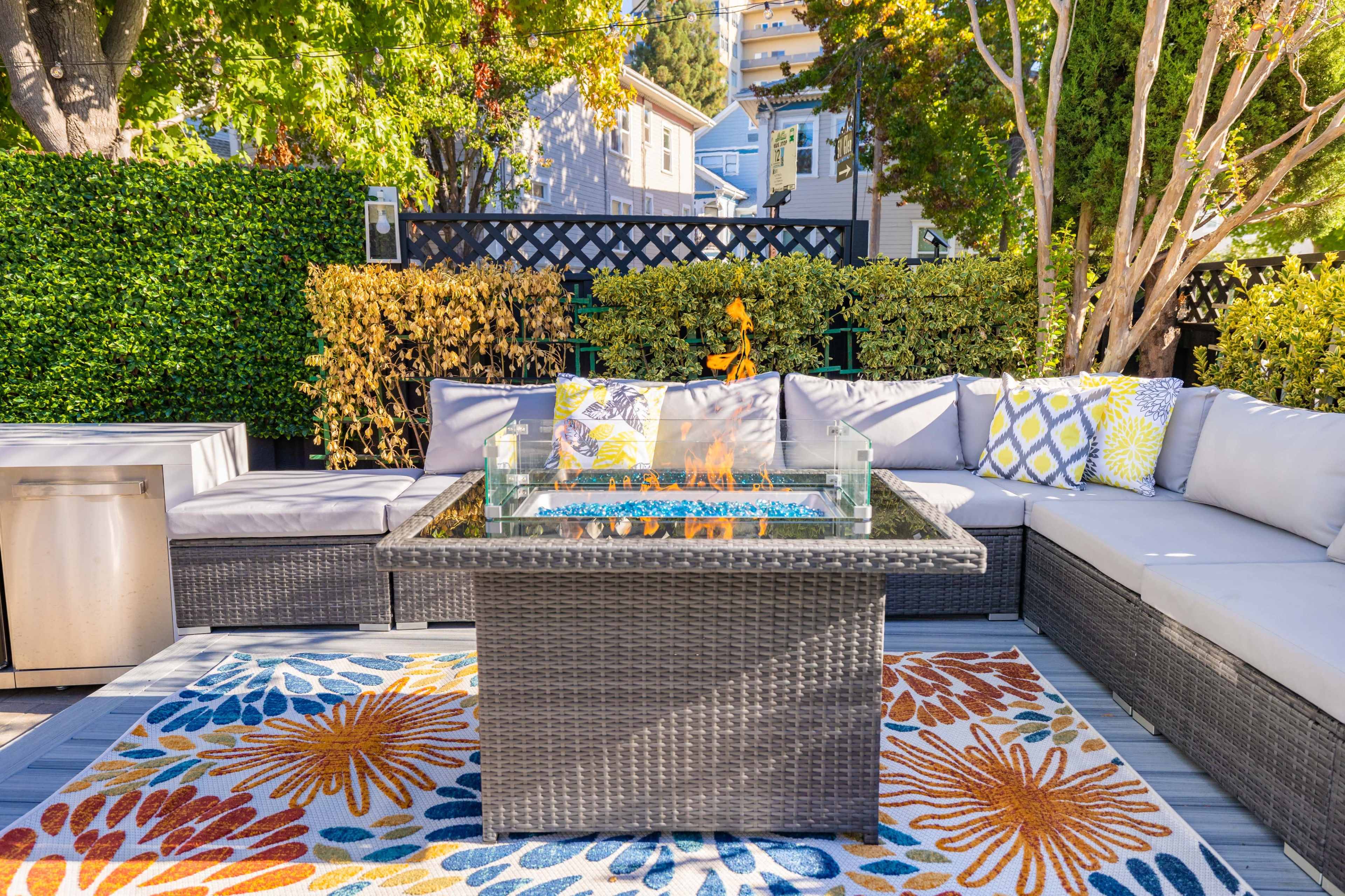 A patio area features a cushioned sectional seating arrangement surrounding a rectangular fire pit adorned with blue glass, set on an area rug with colorful floral patterns.