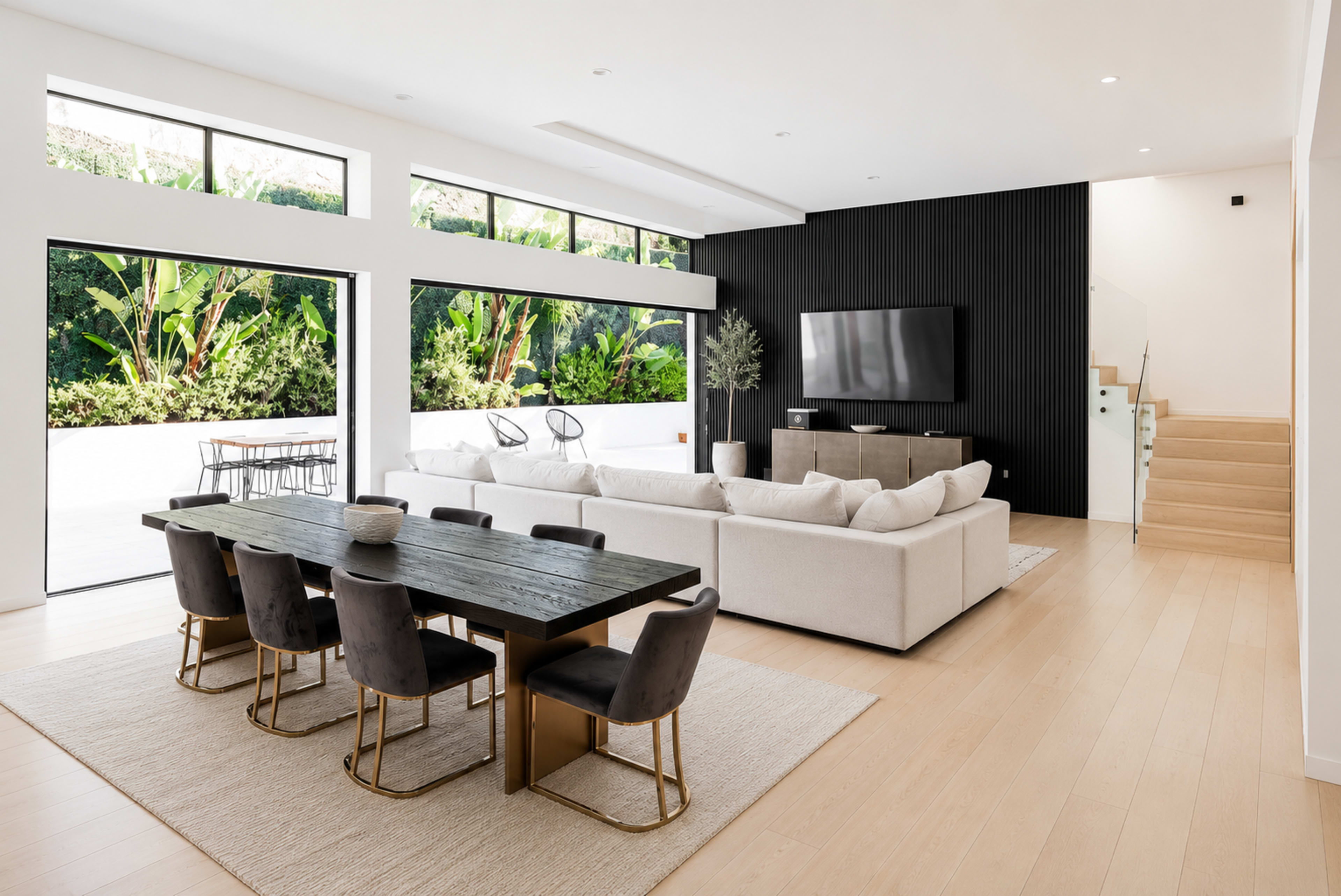 The image shows a modern open-concept living area featuring a dining table with eight chairs, a large sectional sofa, a television mounted on a black wall, and large windows overlooking greenery.