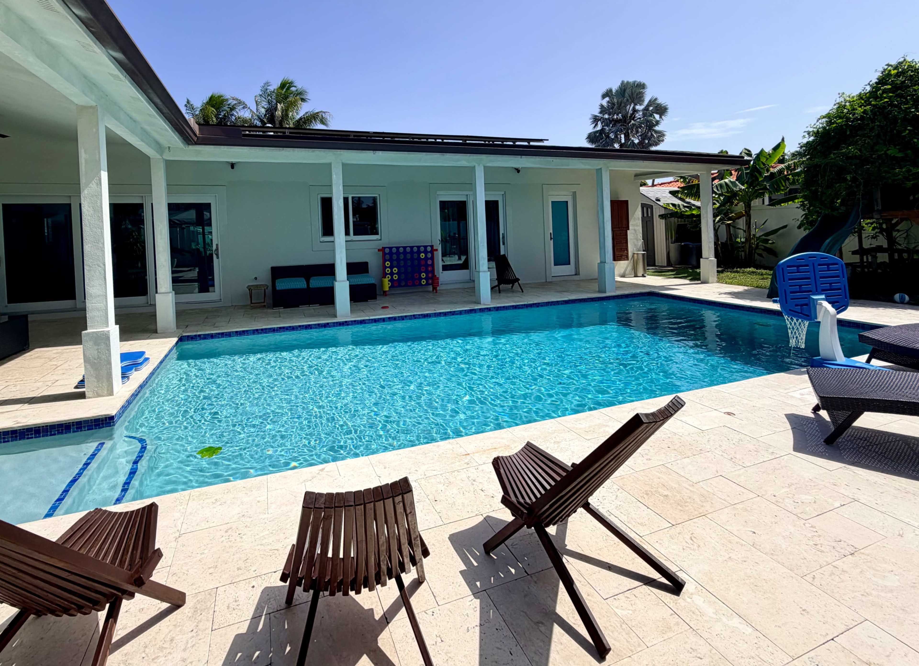 The image shows a backyard swimming pool surrounded by lounge chairs and a modern house in a tropical setting.