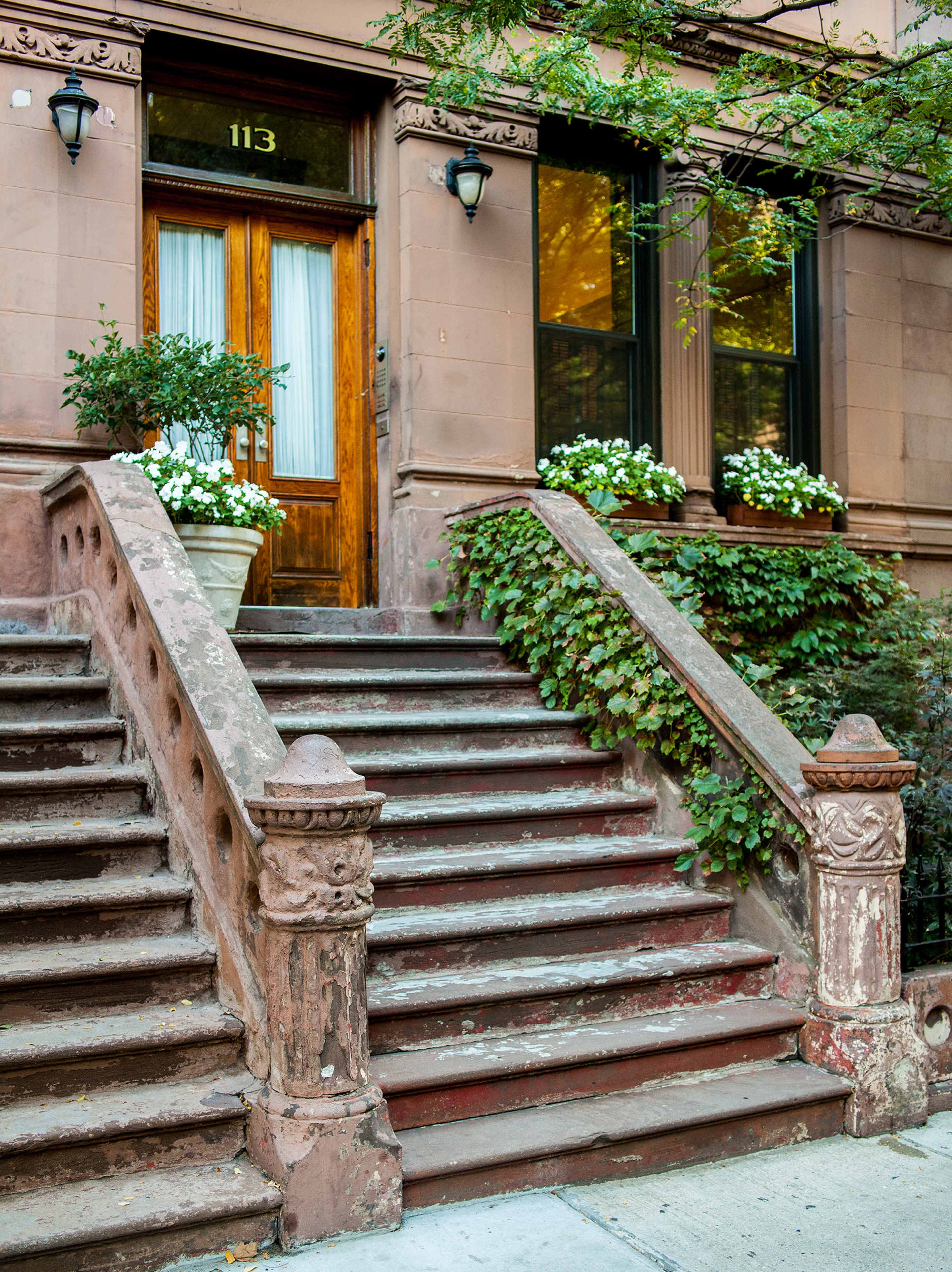 The image shows a brownstone building with a wooden door, flanked by potted plants and steps adorned with ivy and white flowers.