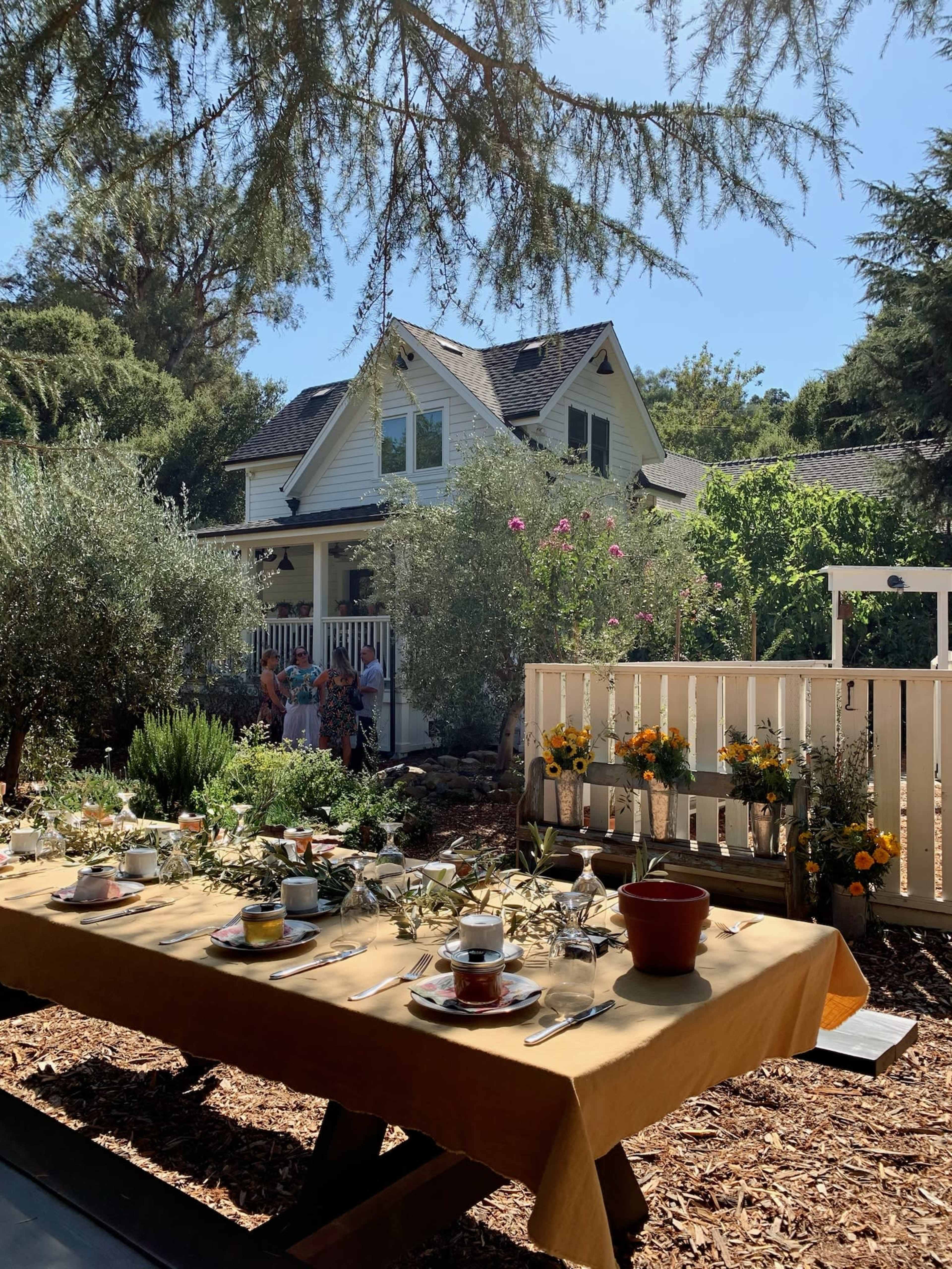 An outdoor table set with dishes and tea cups is positioned in front of a yellow house surrounded by greenery and flowers.