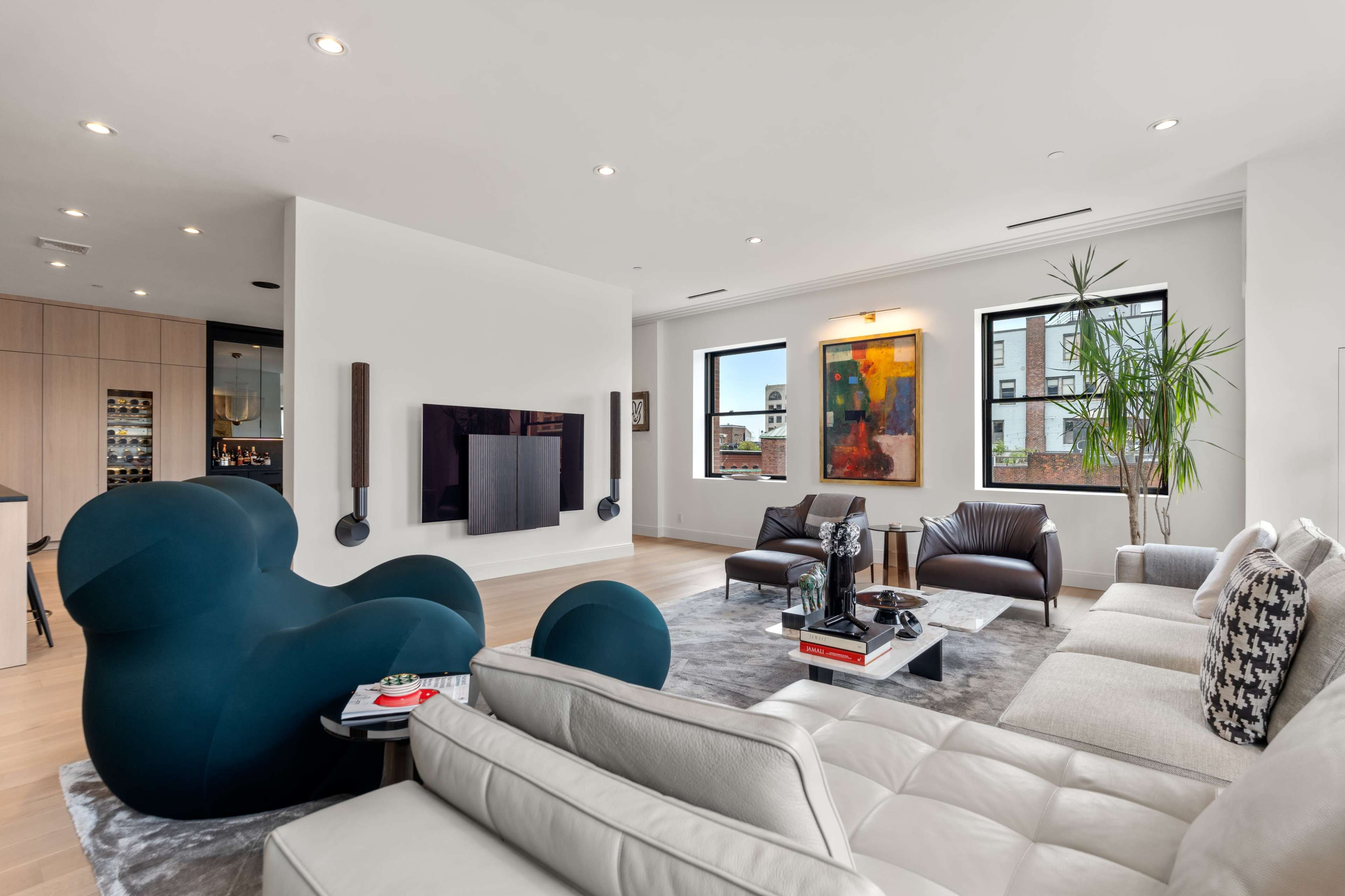 A modern living room features a mix of seating with a curved blue chair, a white leather sofa, and brown leather chairs, along with a large television mounted on the wall and vibrant artwork.