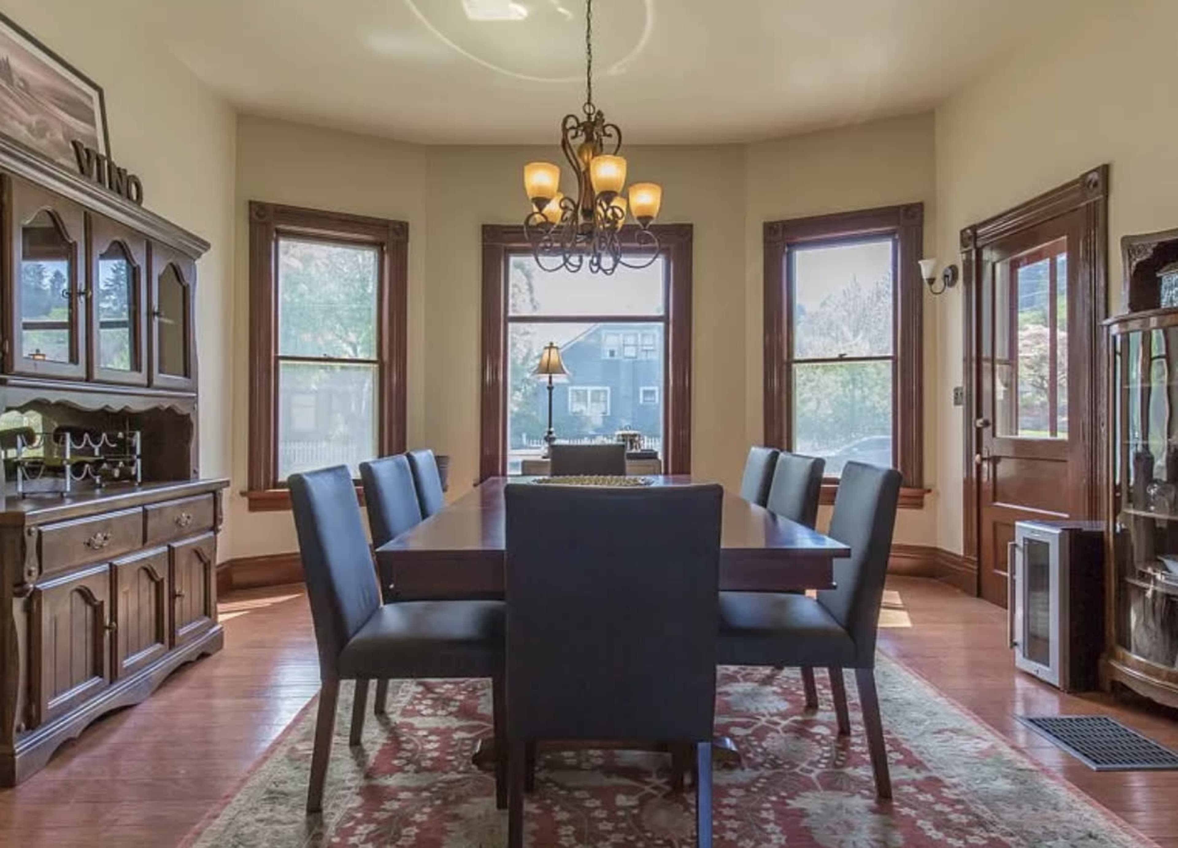 A dining room featuring a long wooden table with six upholstered chairs, surrounded by large windows and wooden furniture.