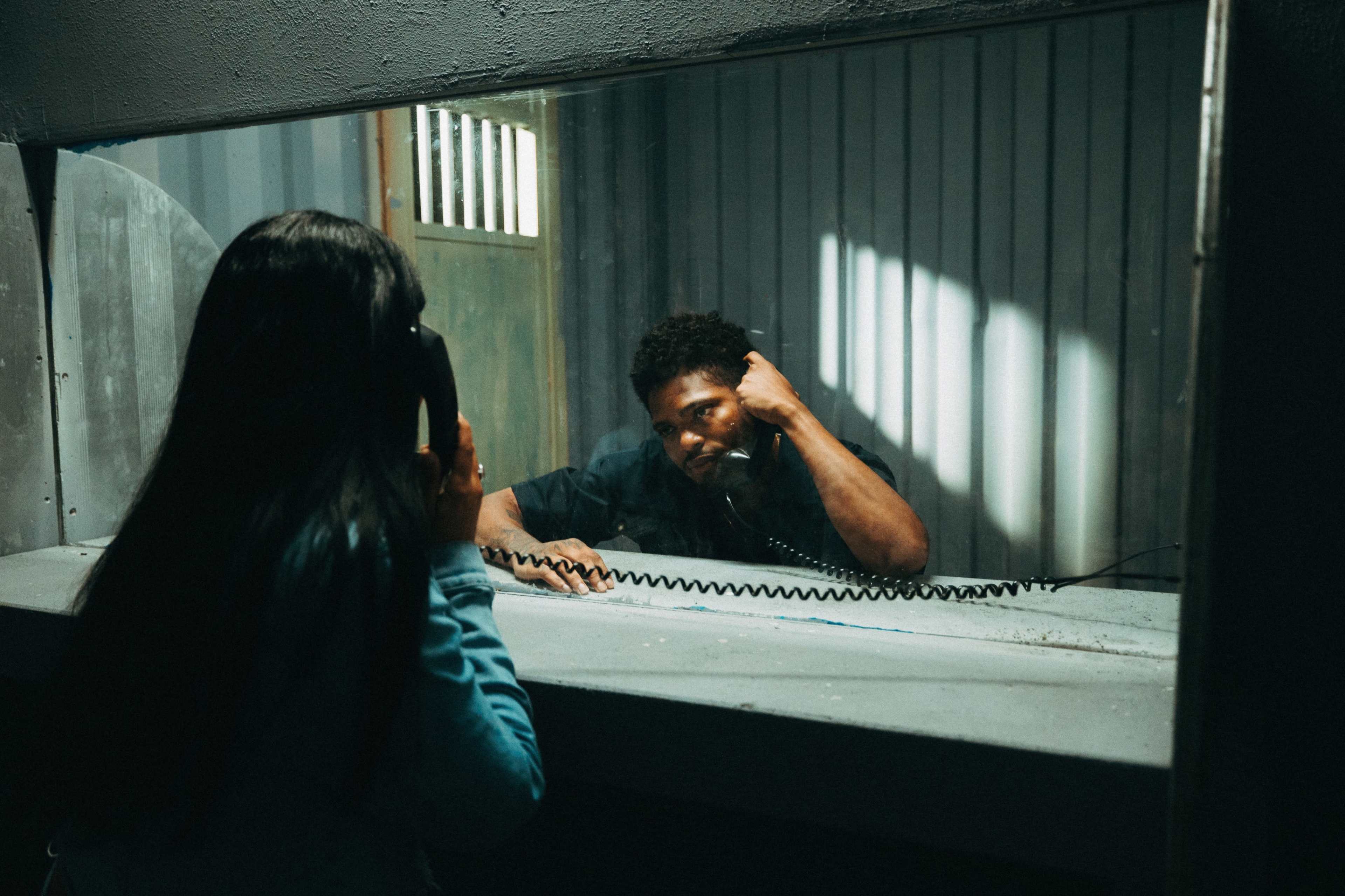 A woman speaks into a payphone while a man, seated behind a glass partition, listens on the other end of the call.