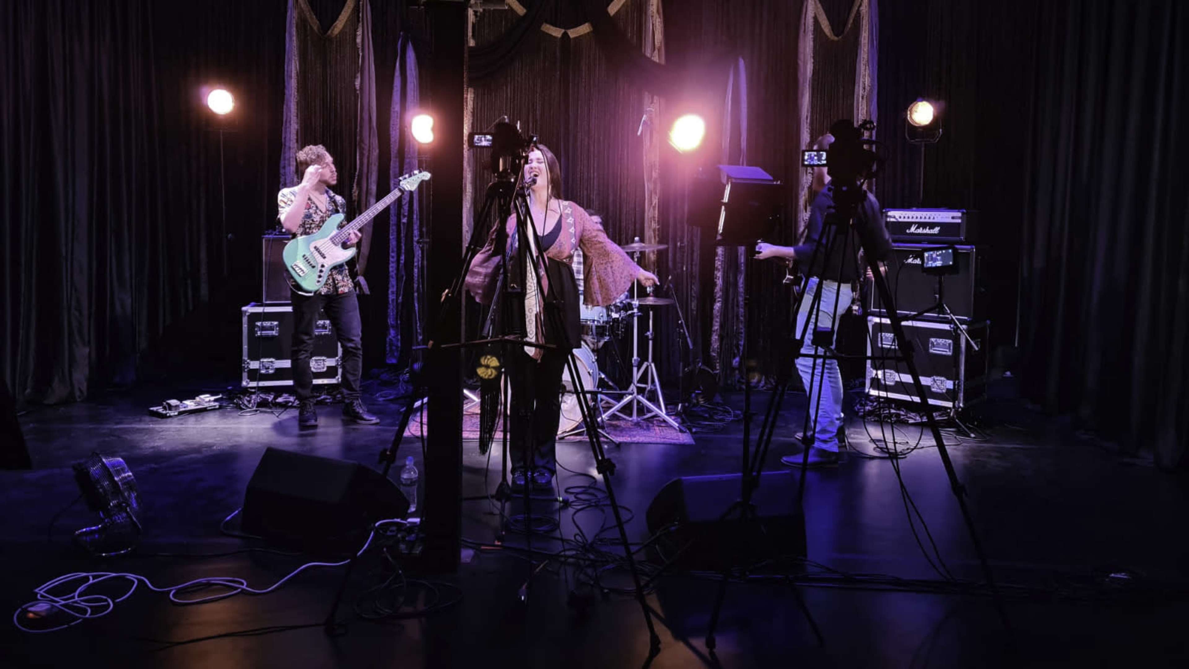 A band performs on stage with musical instruments and camera equipment set up for recording.