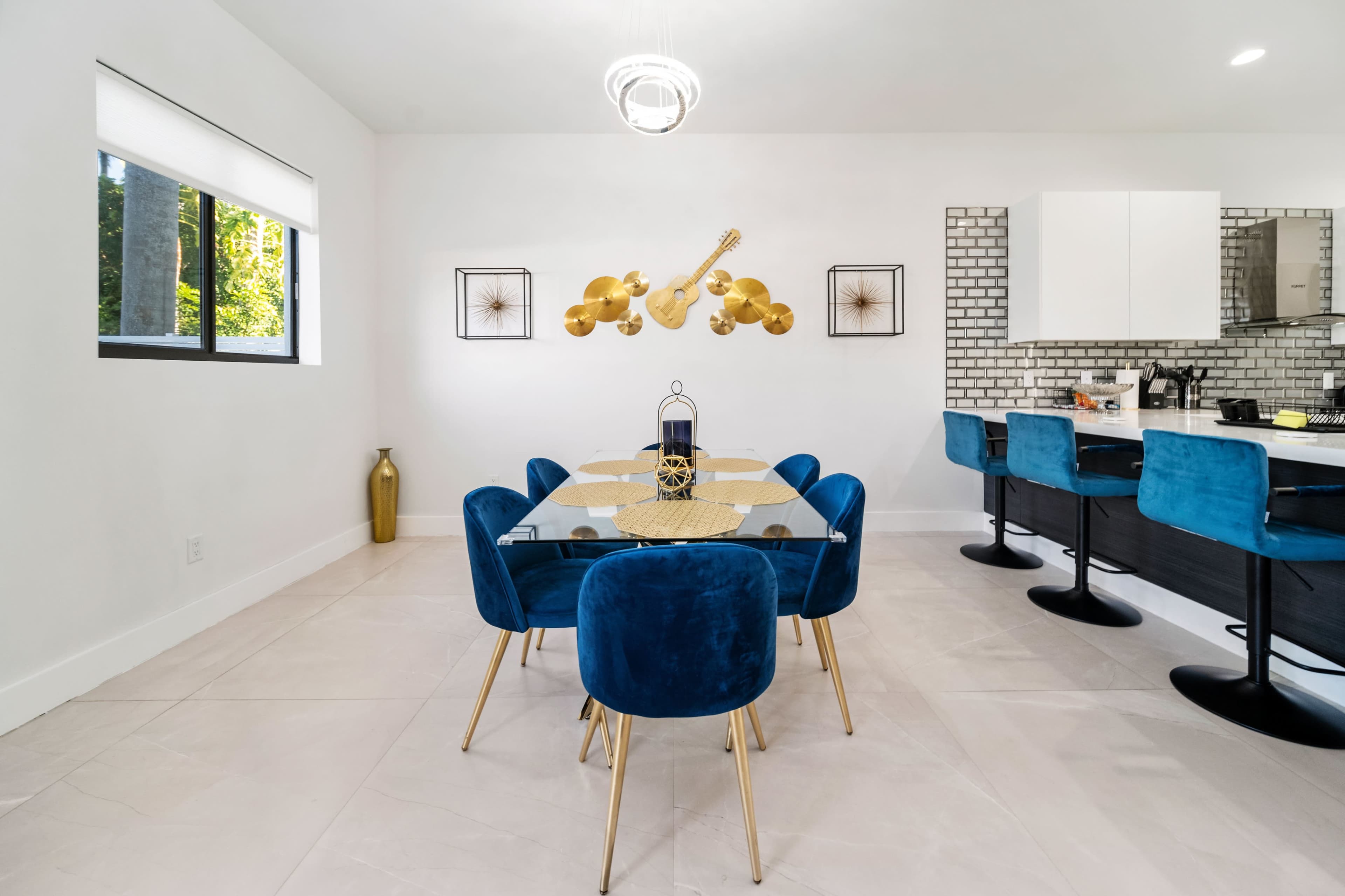 A modern dining area with a glass table surrounded by blue velvet chairs, adjacent to a kitchen featuring white cabinetry and a brick backsplash.