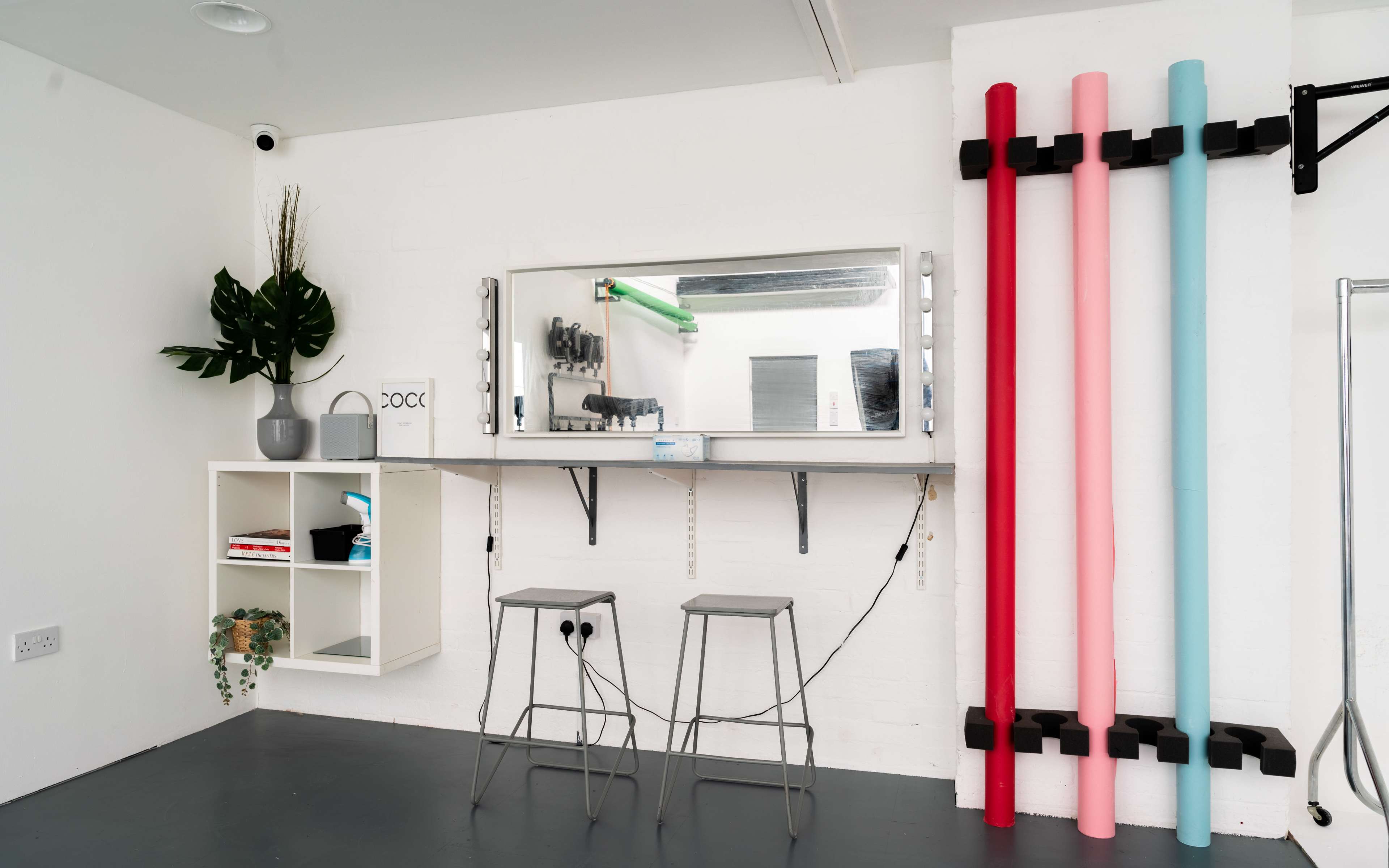 A minimalist room featuring a mirror above a countertop with two stools and colorful pipes mounted on the wall.