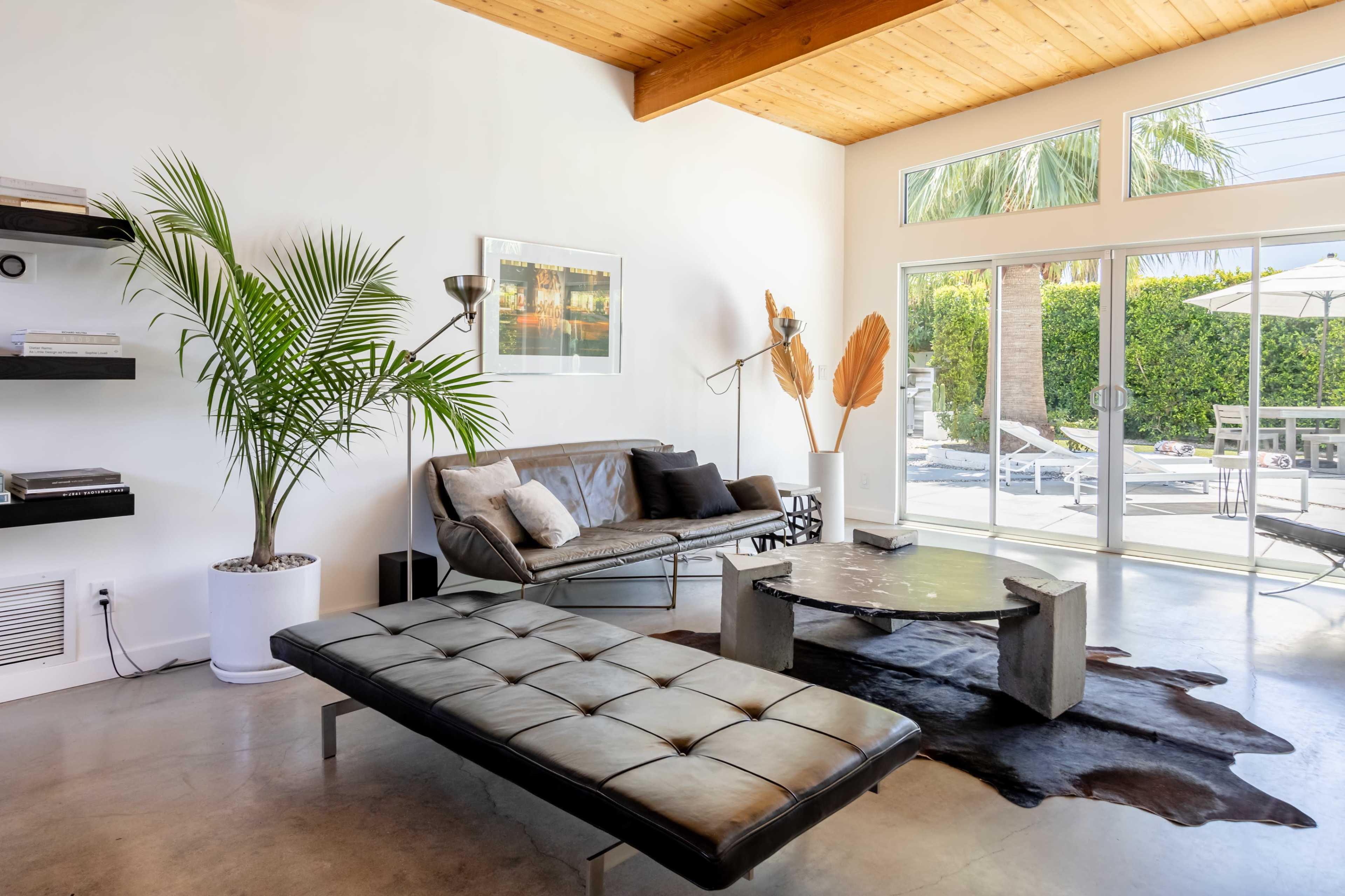 A modern living room features a black leather sofa and a bench, with a large window overlooking a pool area and palm trees.