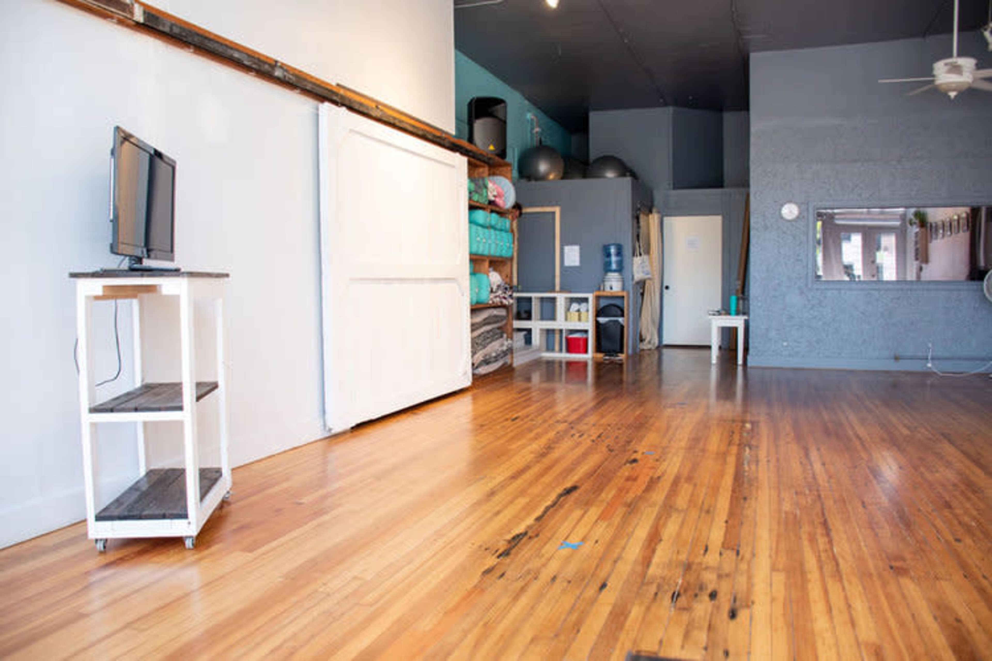 A spacious room with hardwood floors, featuring a television on a cart, storage shelves, and a small table.