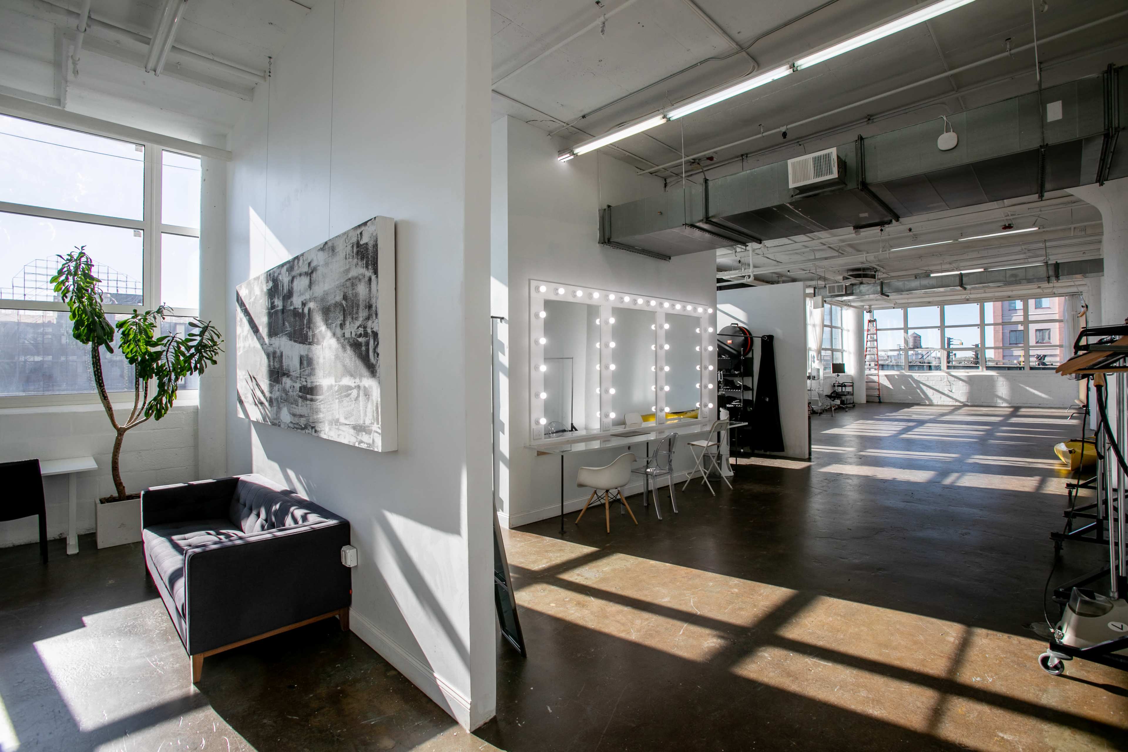 A well-lit studio space featuring a makeup station with bright lights, a seating area, and large windows allowing natural light to enter.