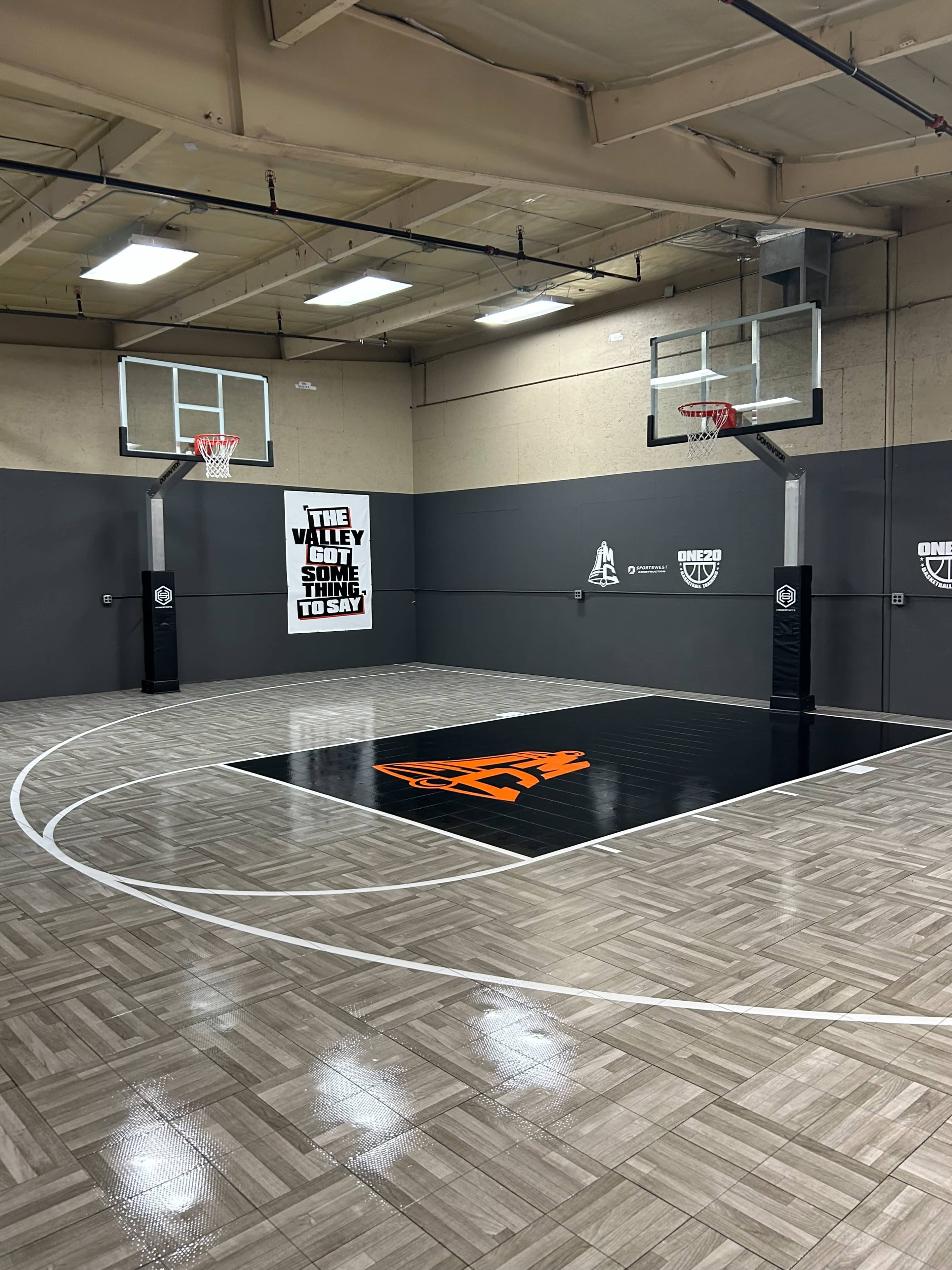 The image shows an indoor basketball court with two hoops, a black center logo, and a light wooden floor.
