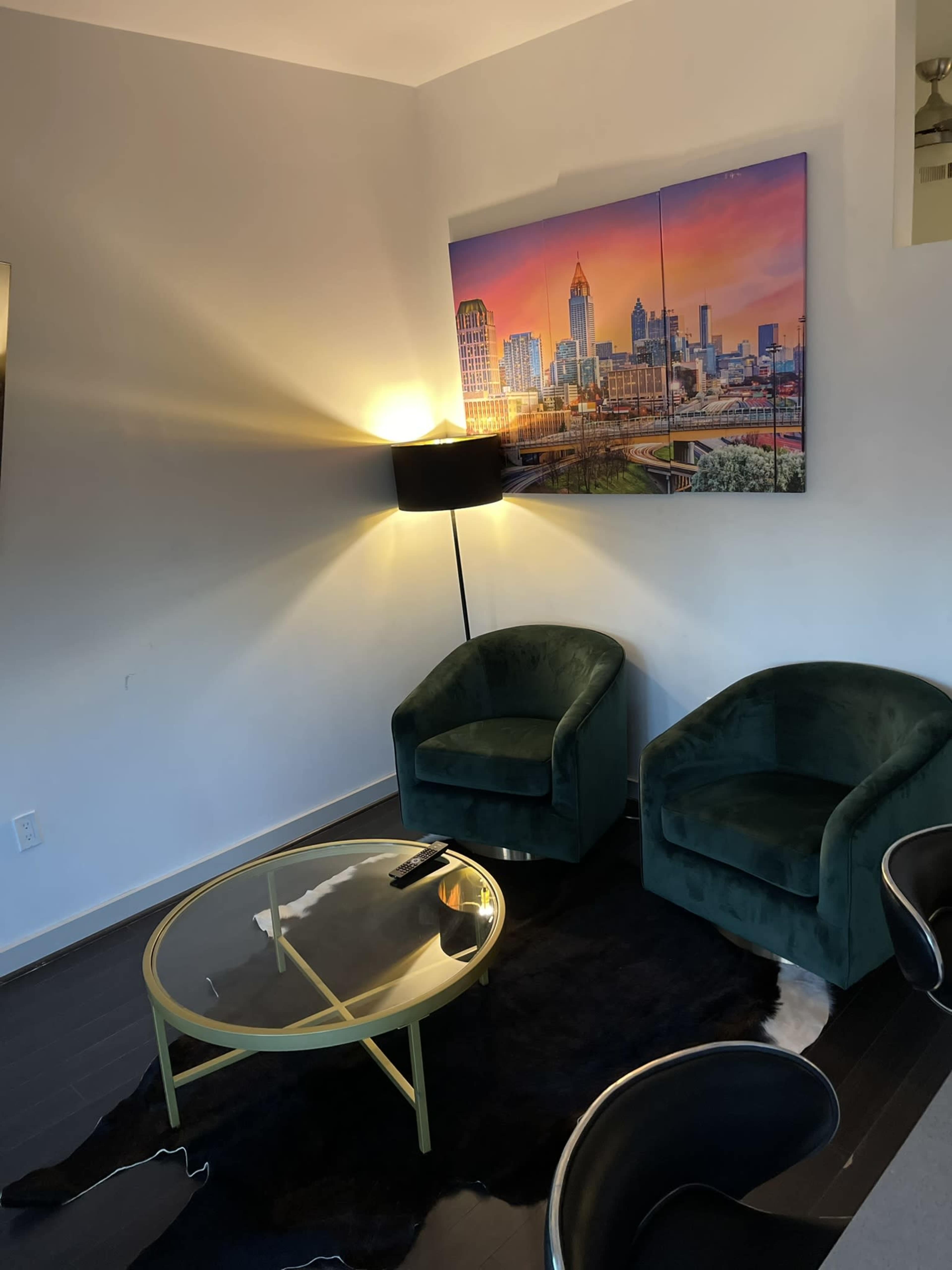 A corner of a living room with two green chairs, a round glass coffee table, and a wall-mounted cityscape painting illuminated by a floor lamp.