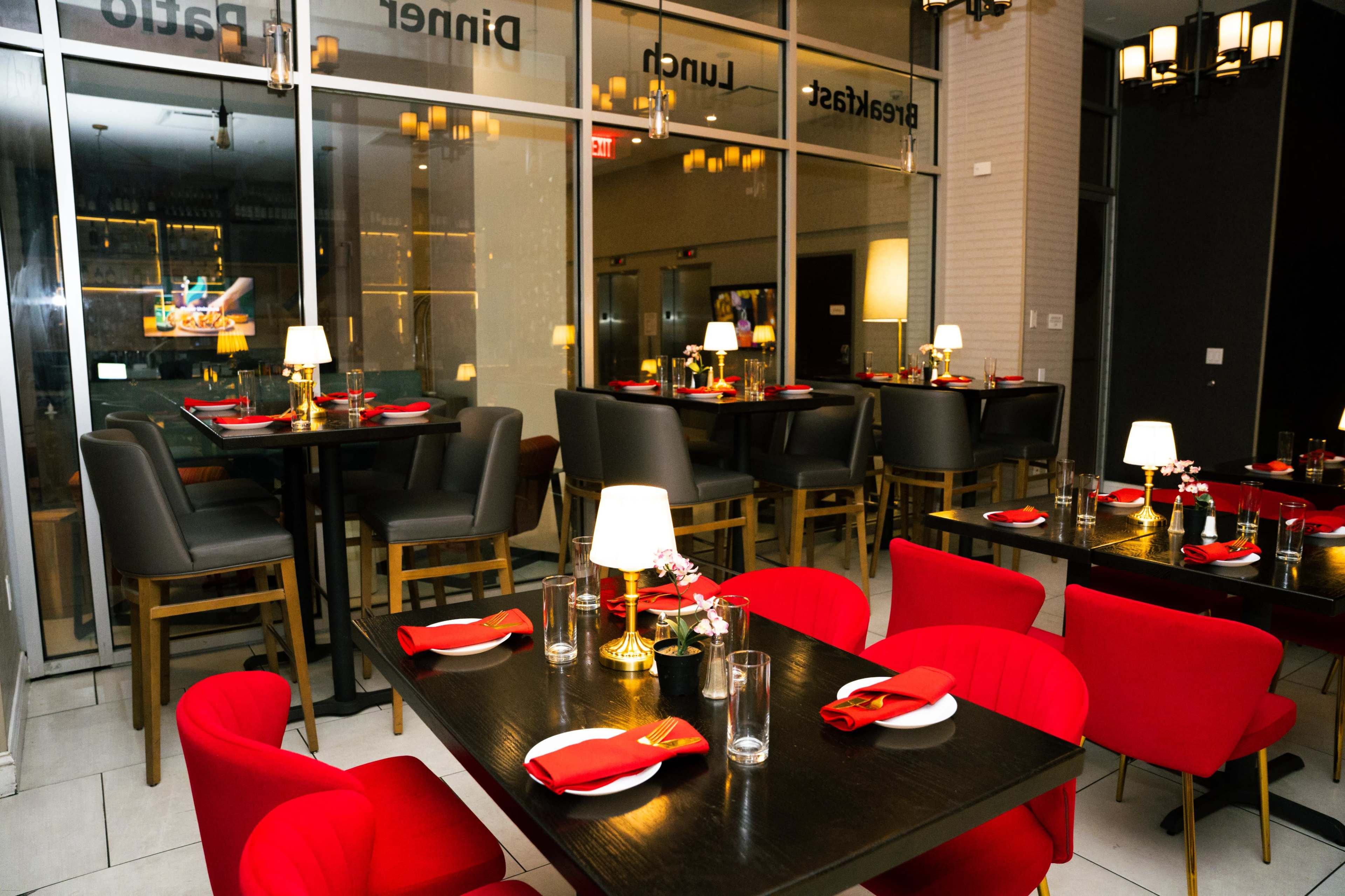 The image shows a modern restaurant interior with red chairs and tables set with white plates and glassware, illuminated by small lamps.