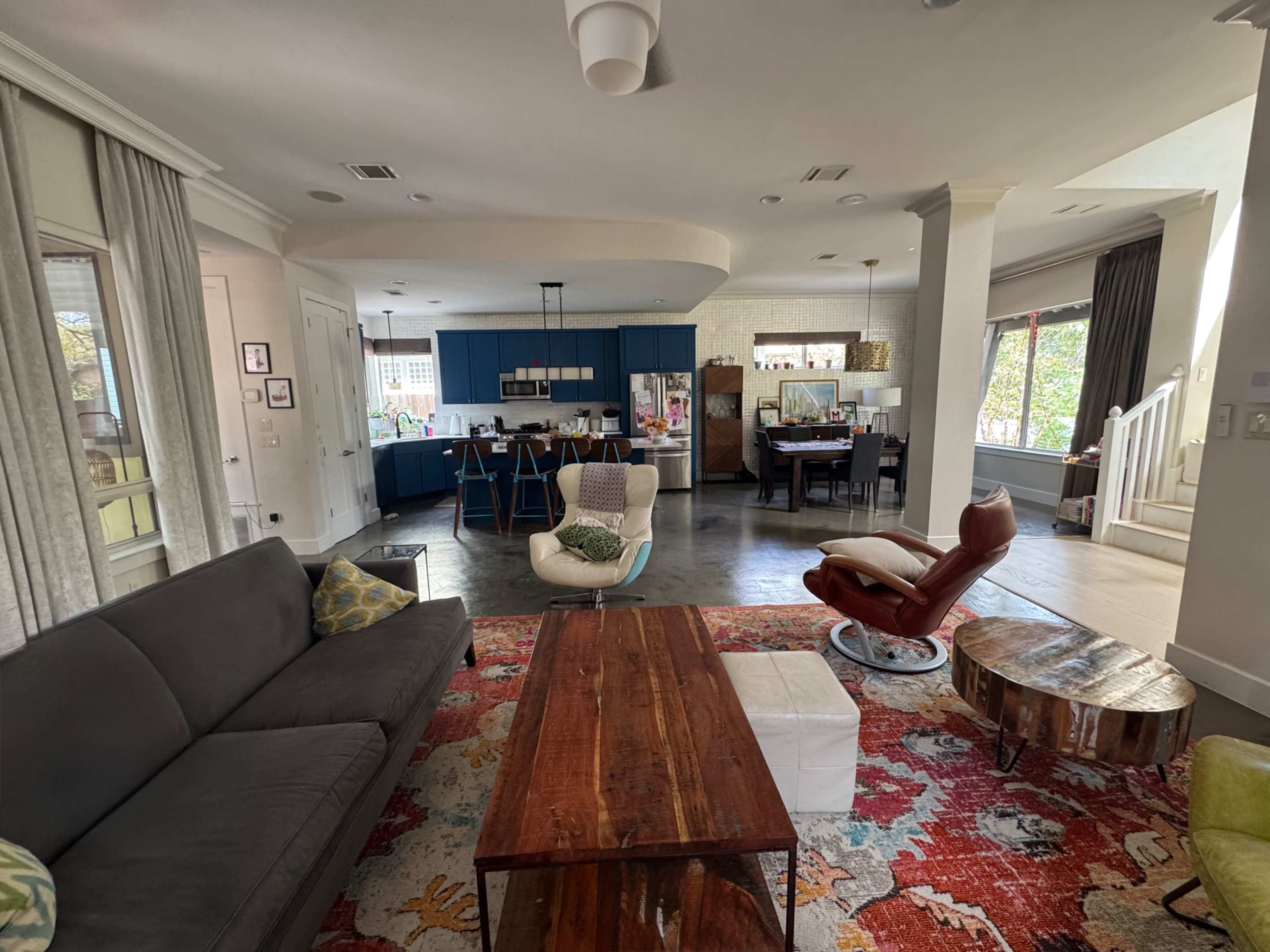 The image shows a spacious, modern living room with a gray couch, a wooden coffee table, and an open kitchen area featuring blue cabinetry and a dining space.