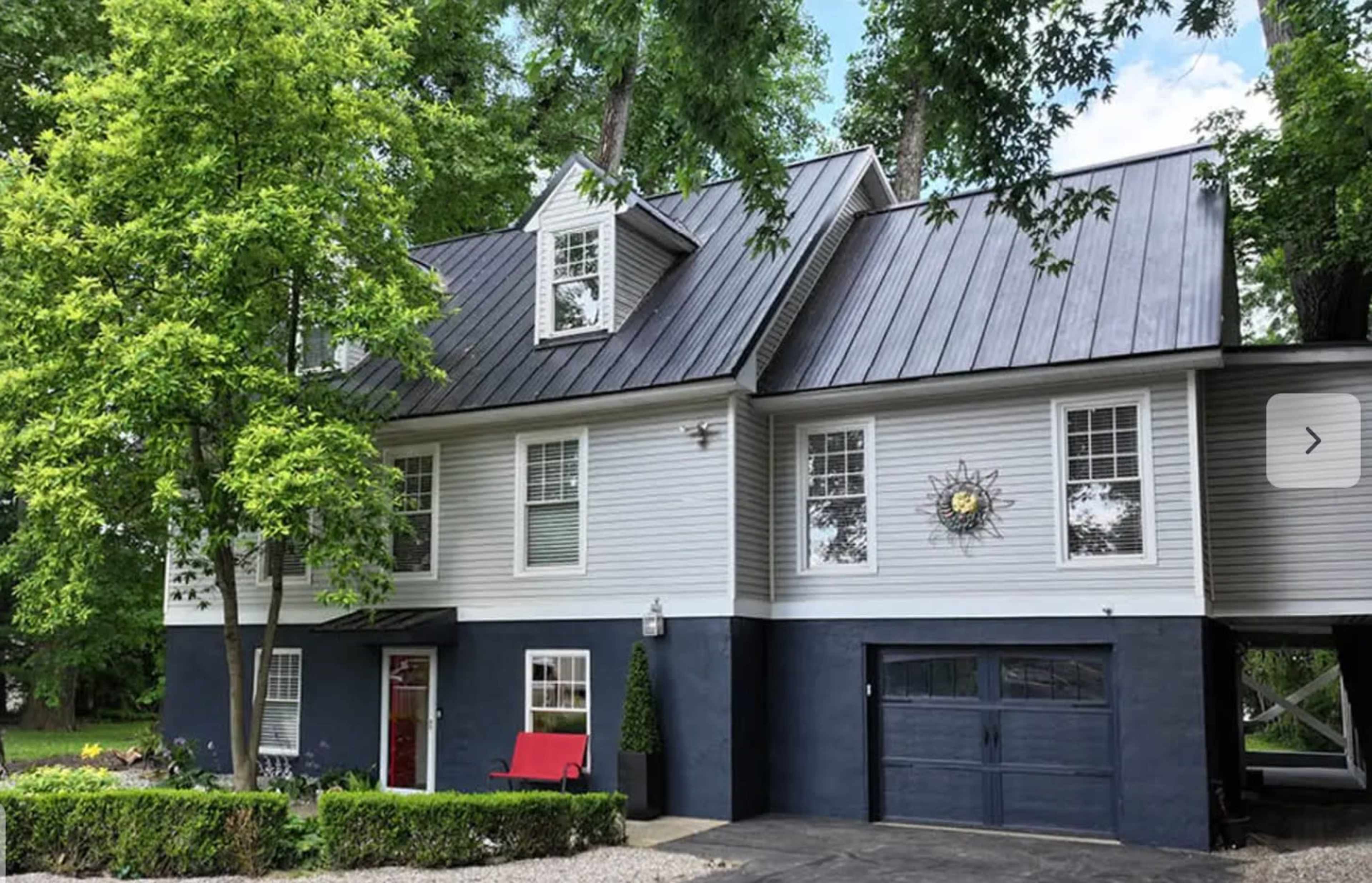 The image shows a two-story house with gray siding, a black metal roof, and a red bench on the porch, surrounded by trees and a landscaped yard.