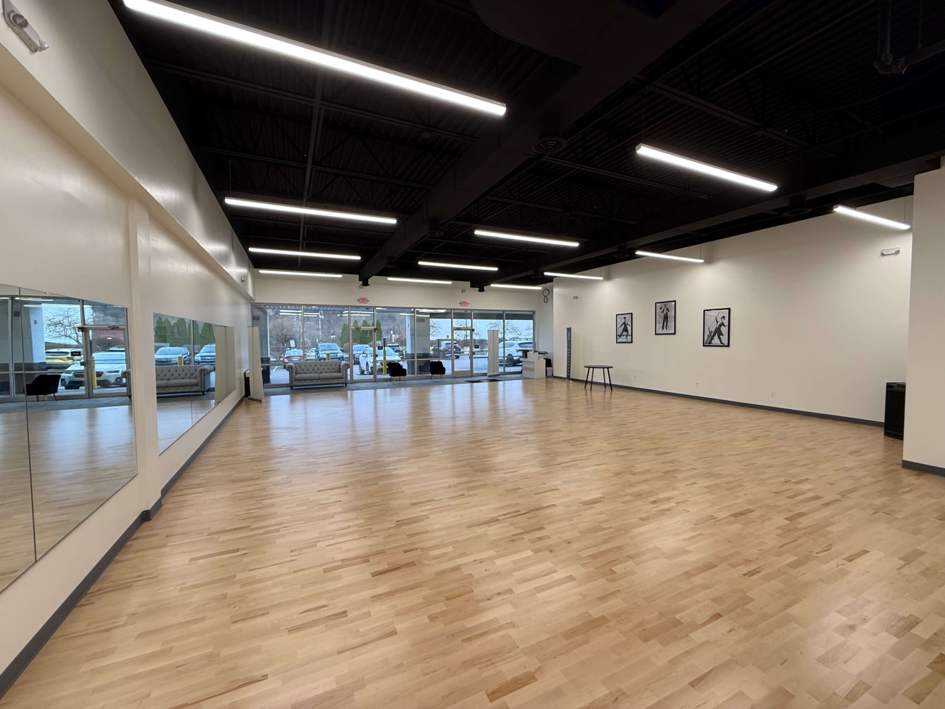 The image shows a spacious, empty room with wooden flooring, large windows at one end, and framed artwork on the walls.