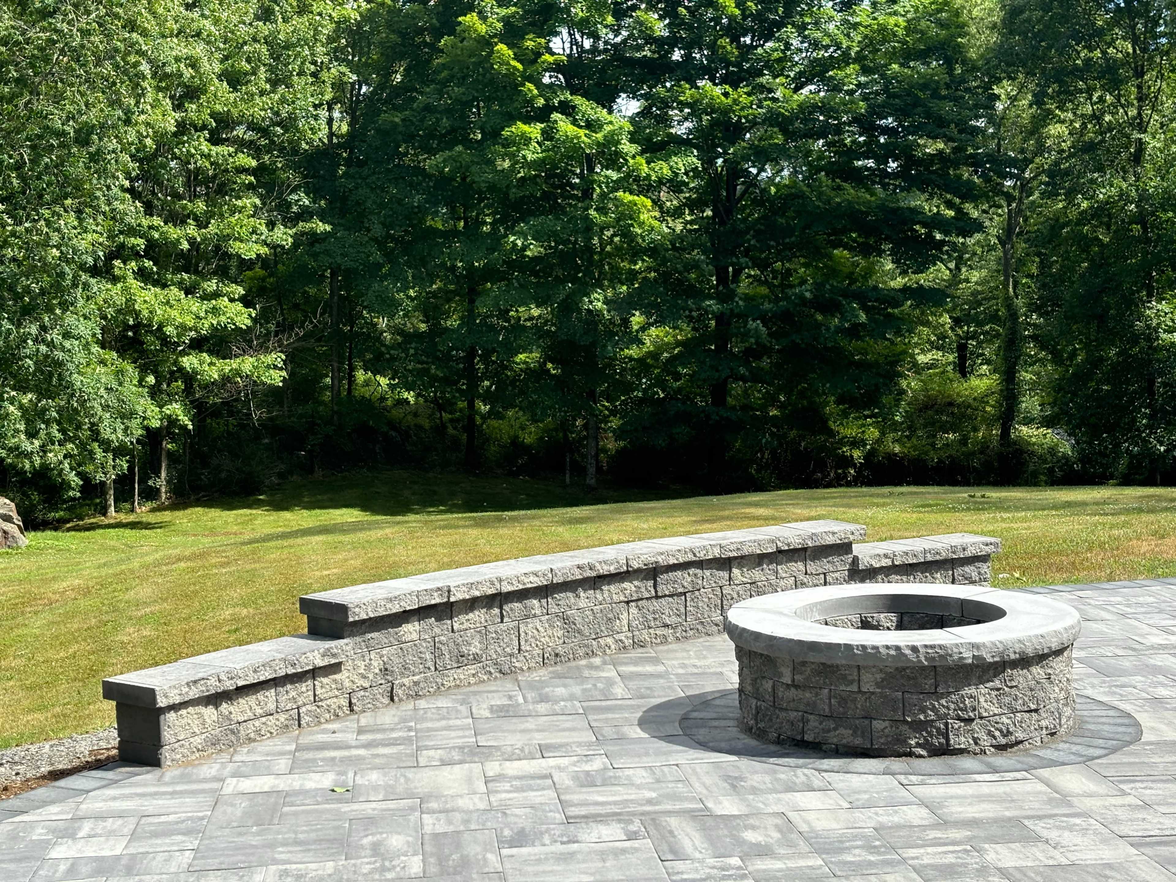 A stone fire pit surrounded by a stone seating area, set in a grassy backyard with trees in the background.