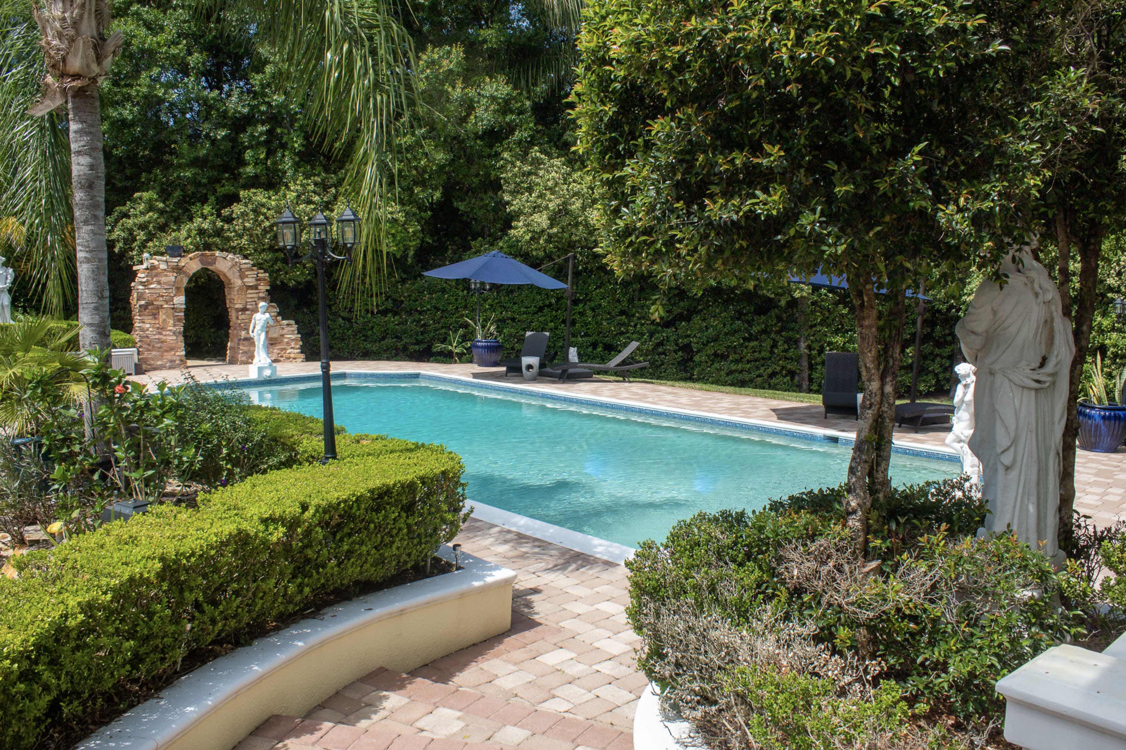 A backyard swimming pool surrounded by greenery, sculptures, and seating areas under blue umbrellas.
