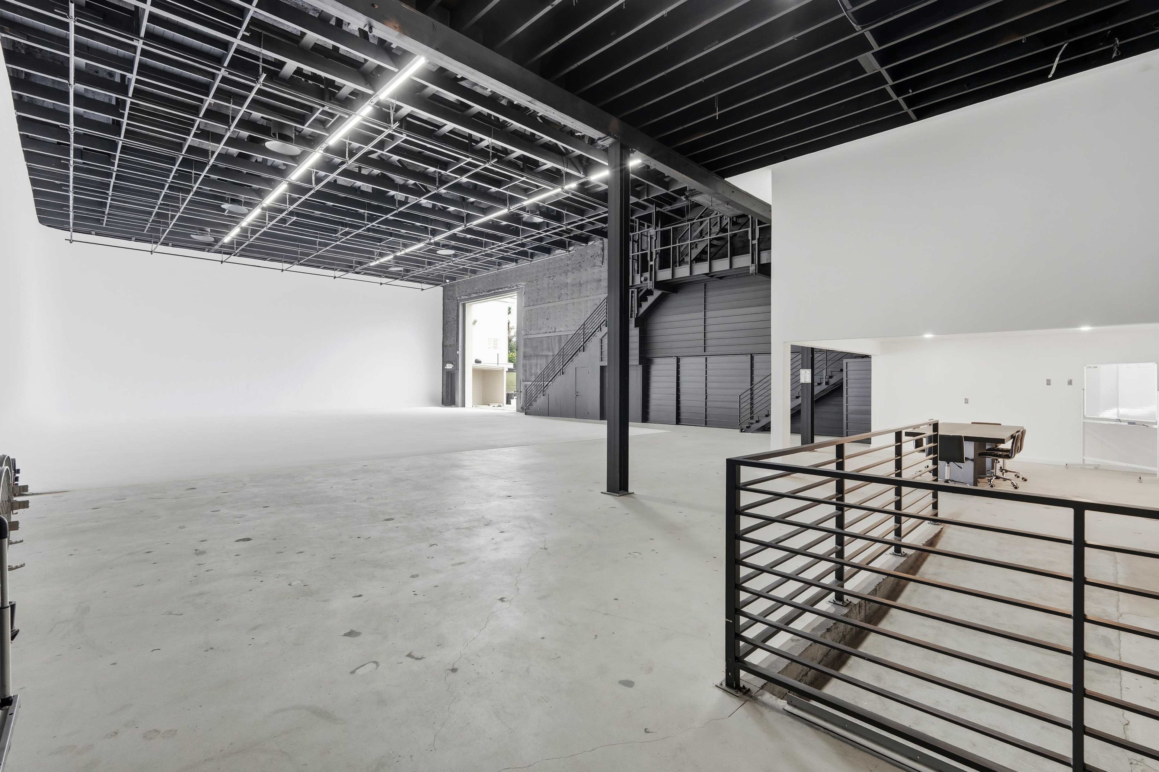 The image shows a spacious, empty studio with a high ceiling, large white walls, and a staircase leading to an upper level.