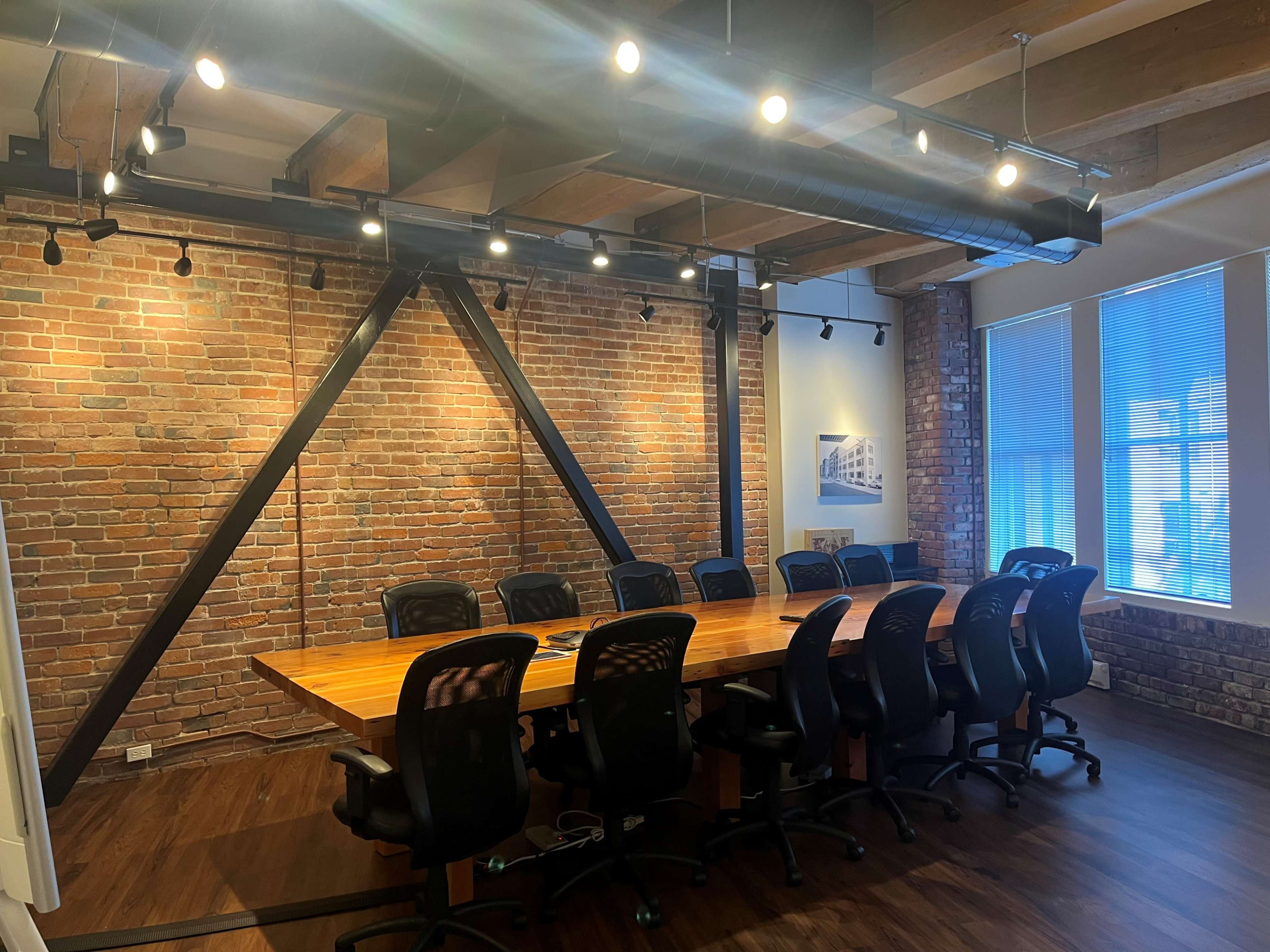 A large conference table with black office chairs is situated in a room featuring exposed brick walls and overhead lighting.