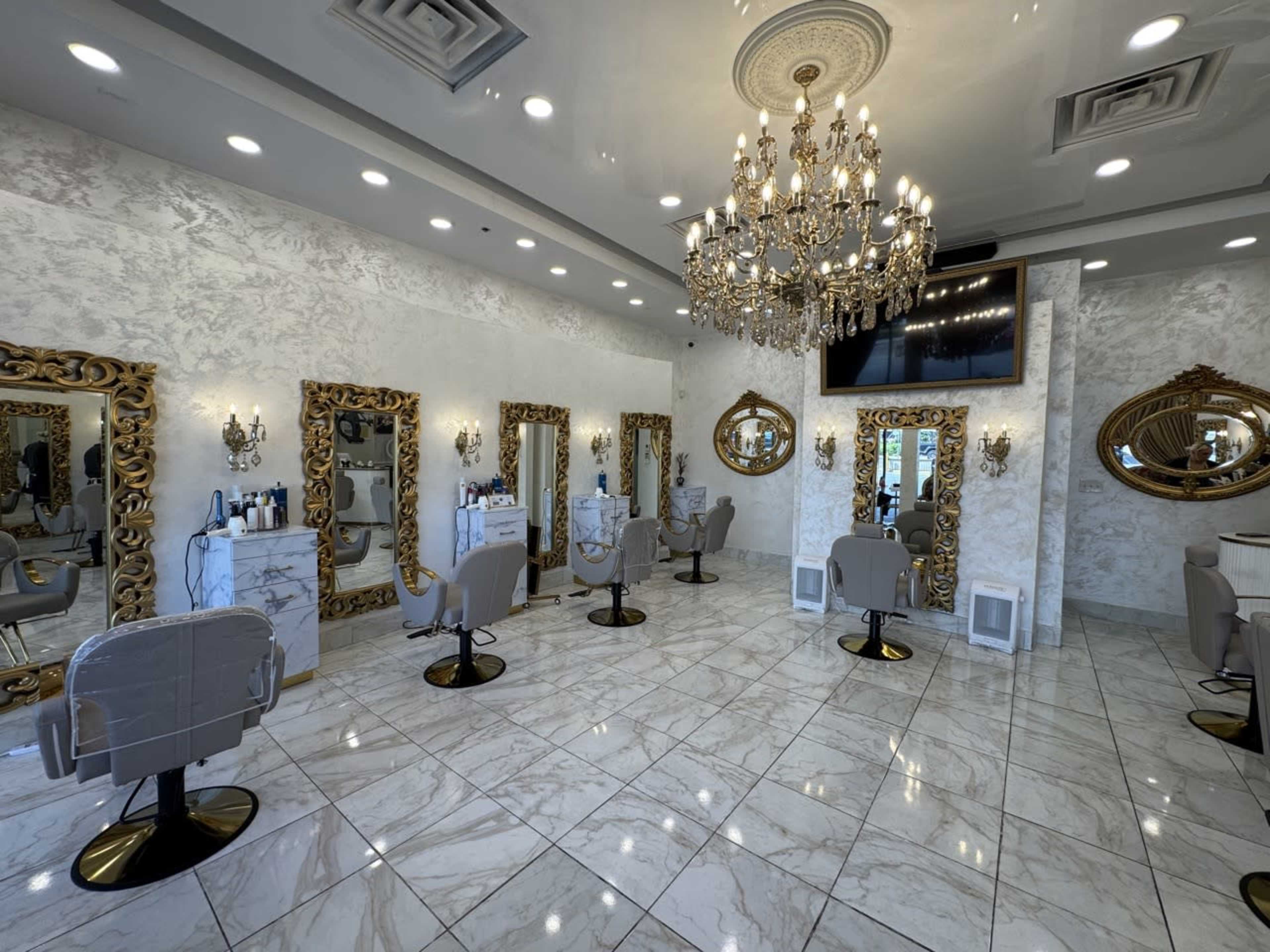 The image shows a brightly lit salon featuring multiple styling stations, each with ornate mirrors and comfortable chairs, decorated with elegant chandeliers and marble flooring.