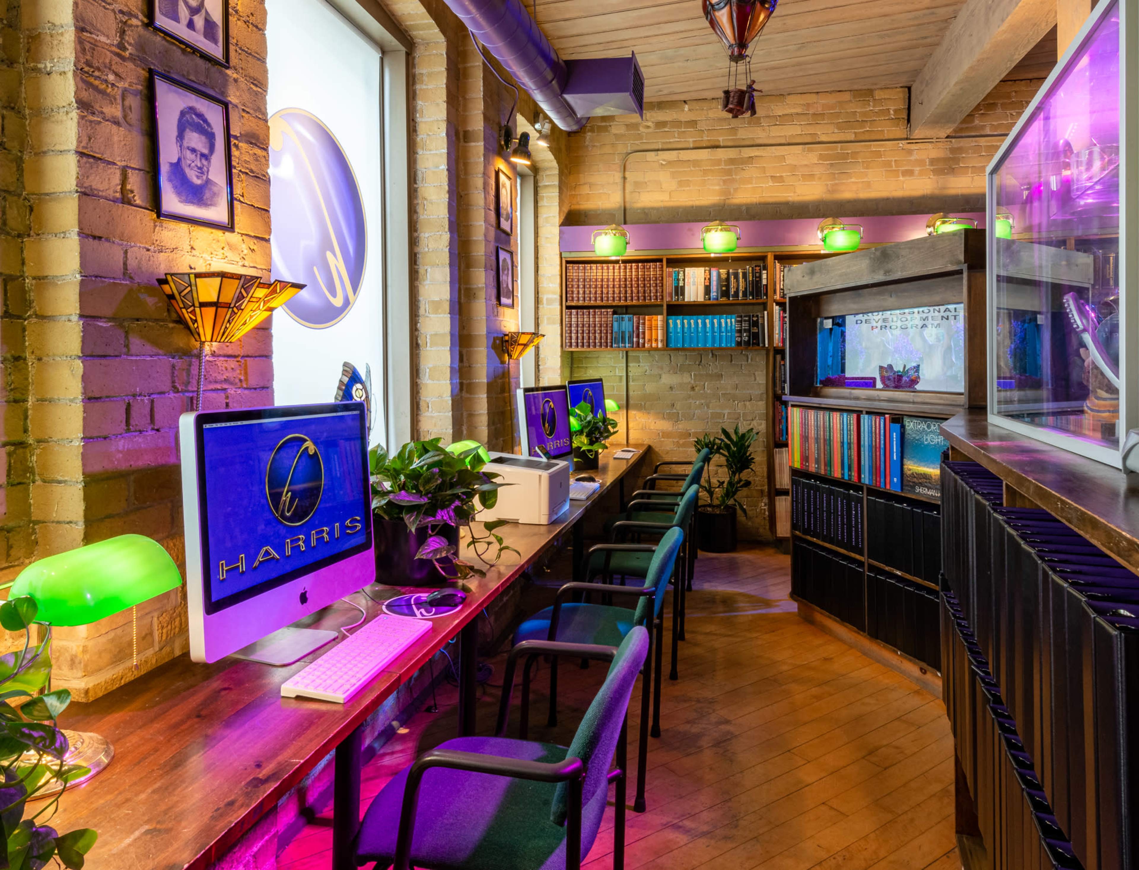 A cozy library-like workspace with computers, bookshelves, and plants, illuminated by colorful lamps.