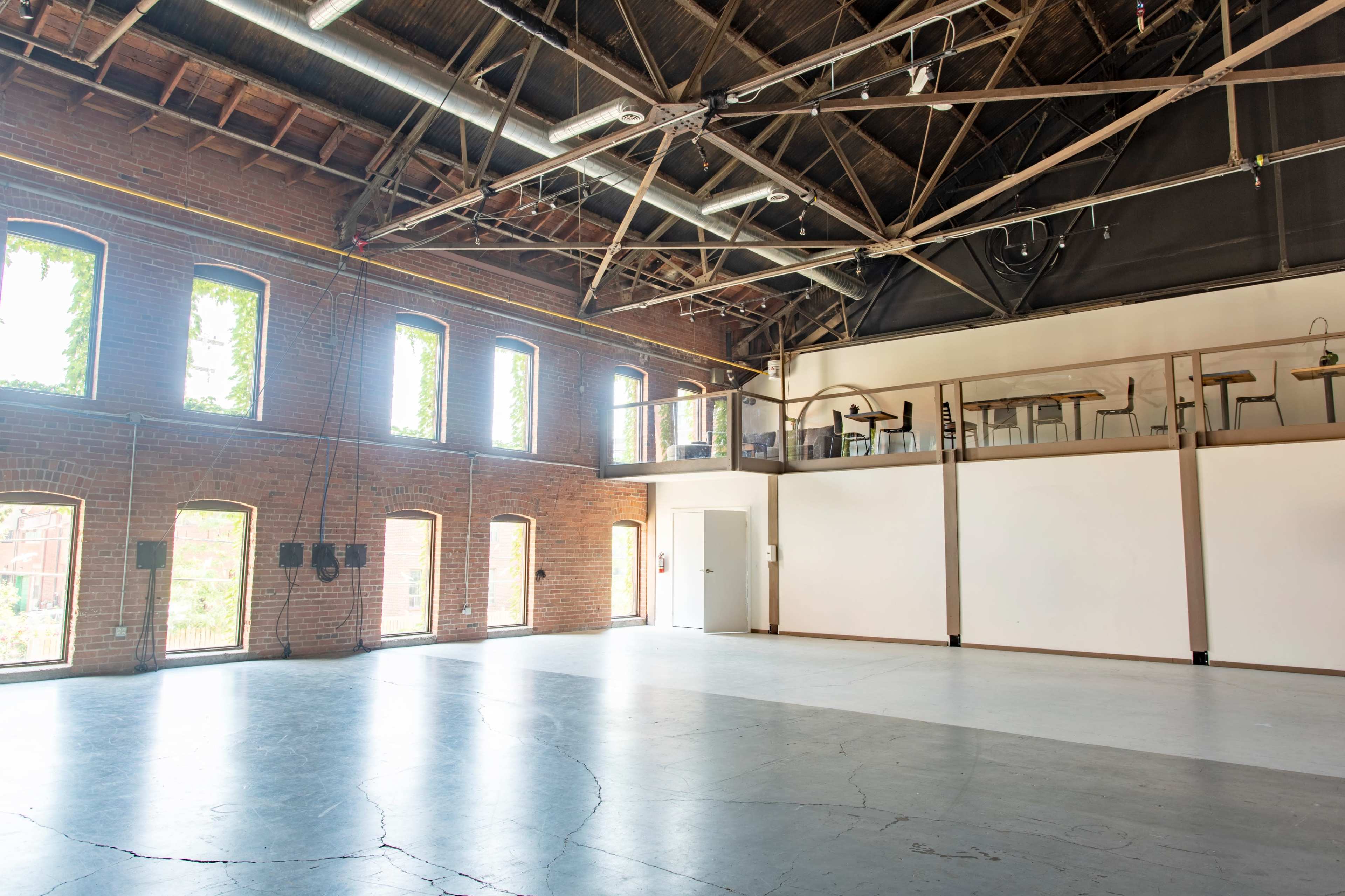 An empty industrial space with tall brick walls, large windows, and a lofted area overlooking the main floor.