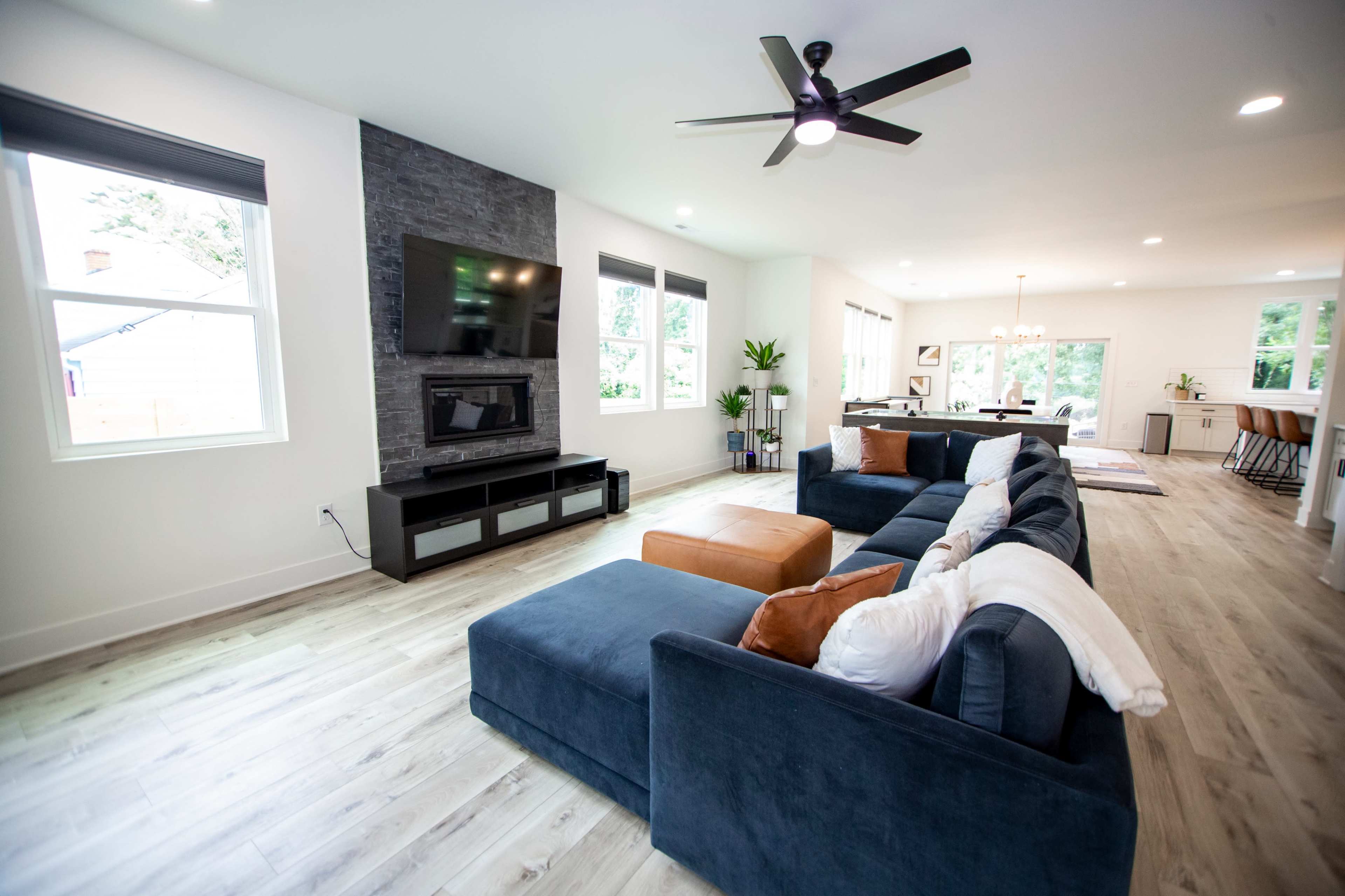 The image shows a modern living room featuring a large blue sectional sofa, a flat-screen TV mounted on a stone wall, and large windows that allow natural light to fill the space.