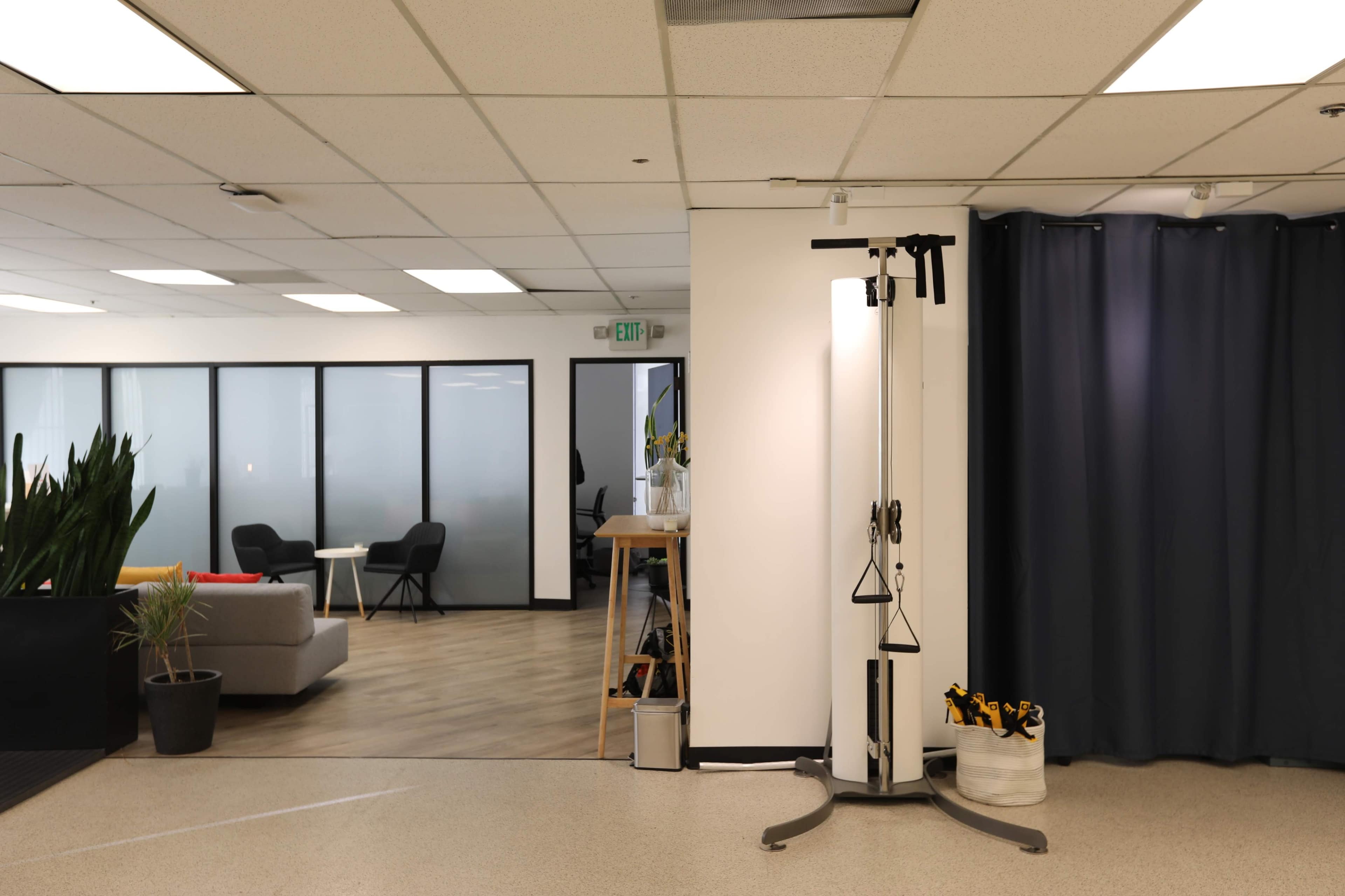 An office space divided by a curtain, with a fitness pulley machine on one side and a seating area visible through glass partitioning on the other.