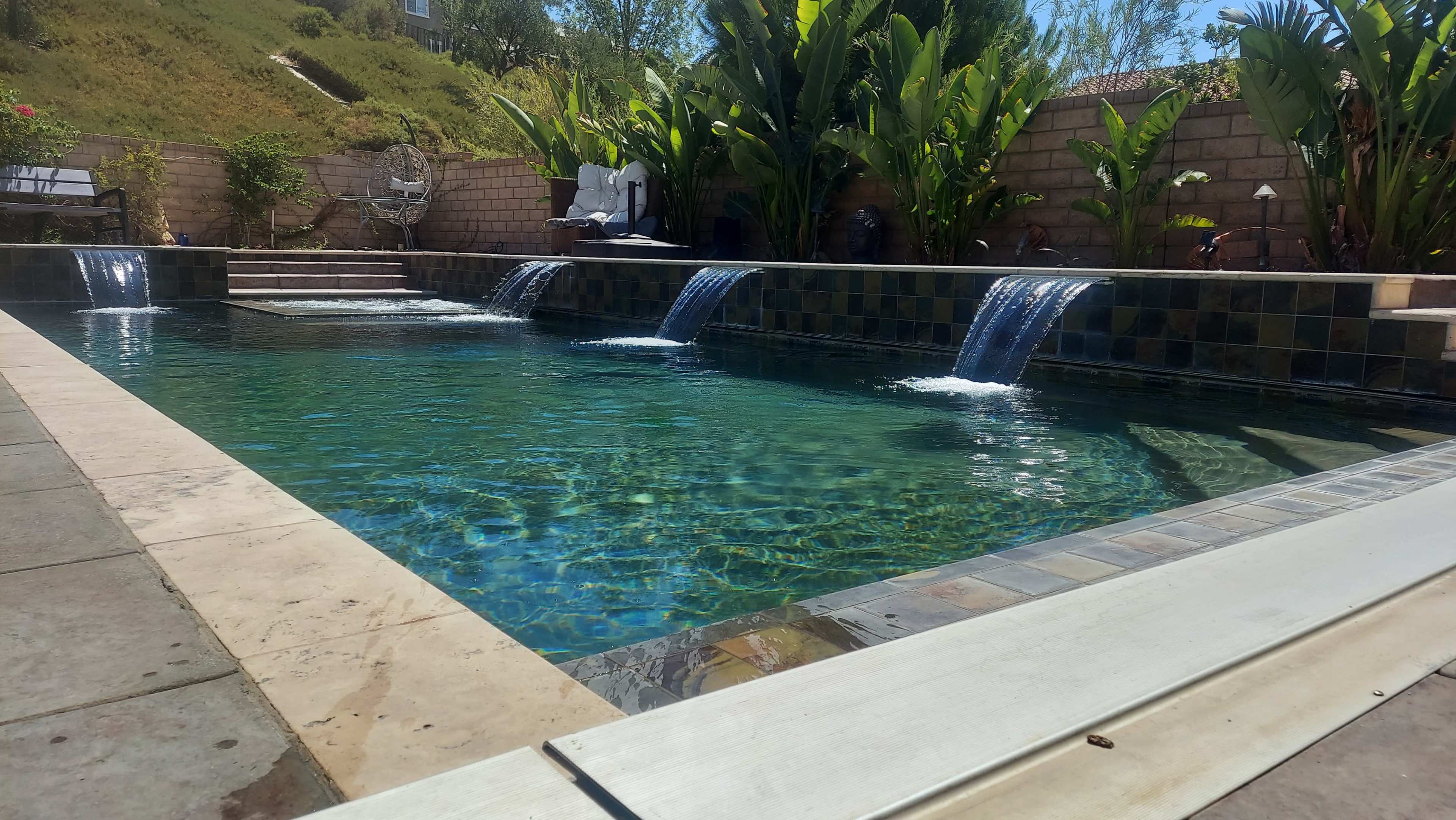 The image shows a swimming pool with three water features spilling into it, surrounded by lush greenery and patio furniture.