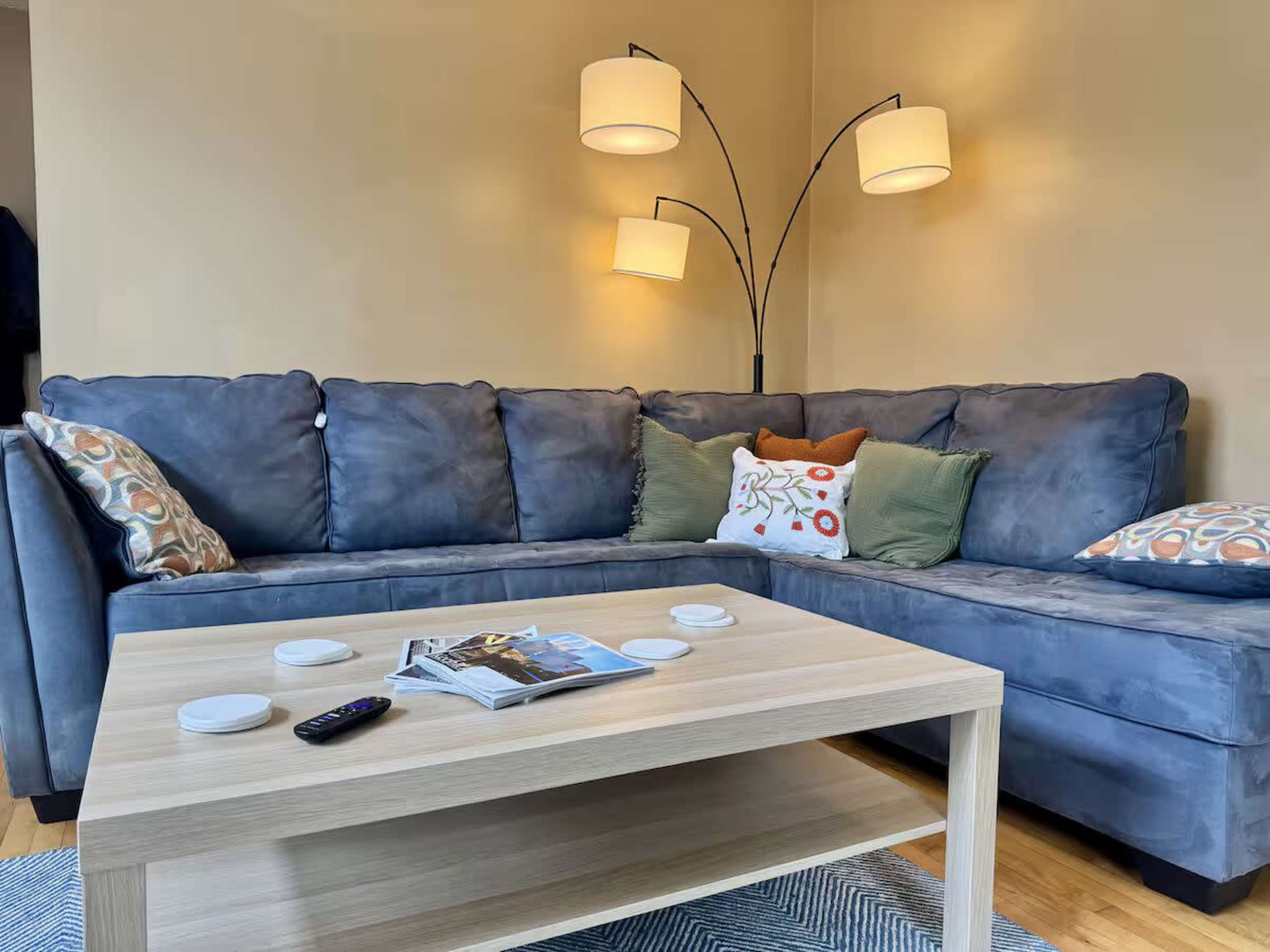 A dark blue sectional couch with decorative pillows is positioned next to a wooden coffee table, which has magazines and a remote, beneath a floor lamp with three adjustable arms.