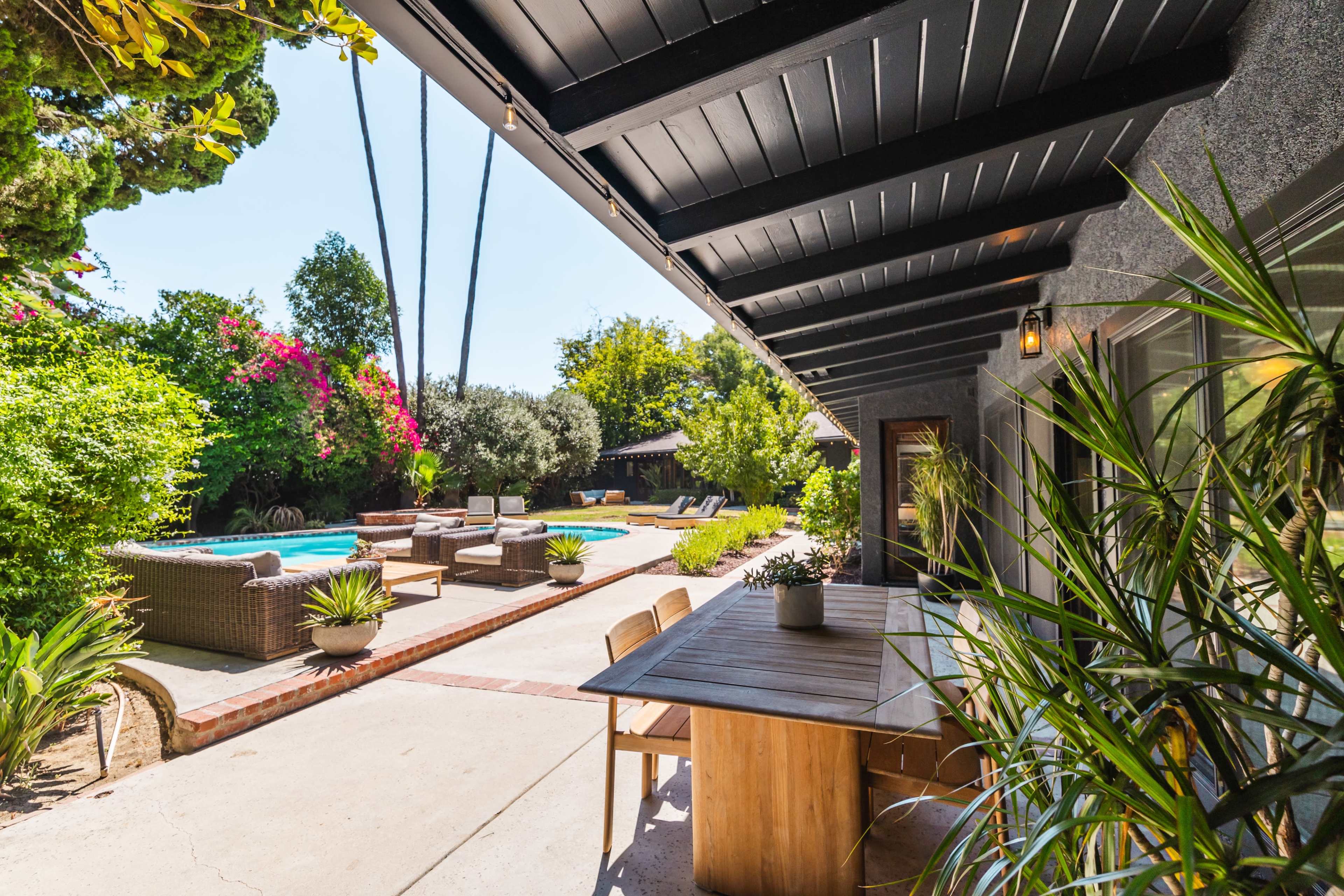 A modern patio area features a wooden table and chairs, surrounded by potted plants, with a view of a swimming pool and lounge chairs in a lush garden setting.