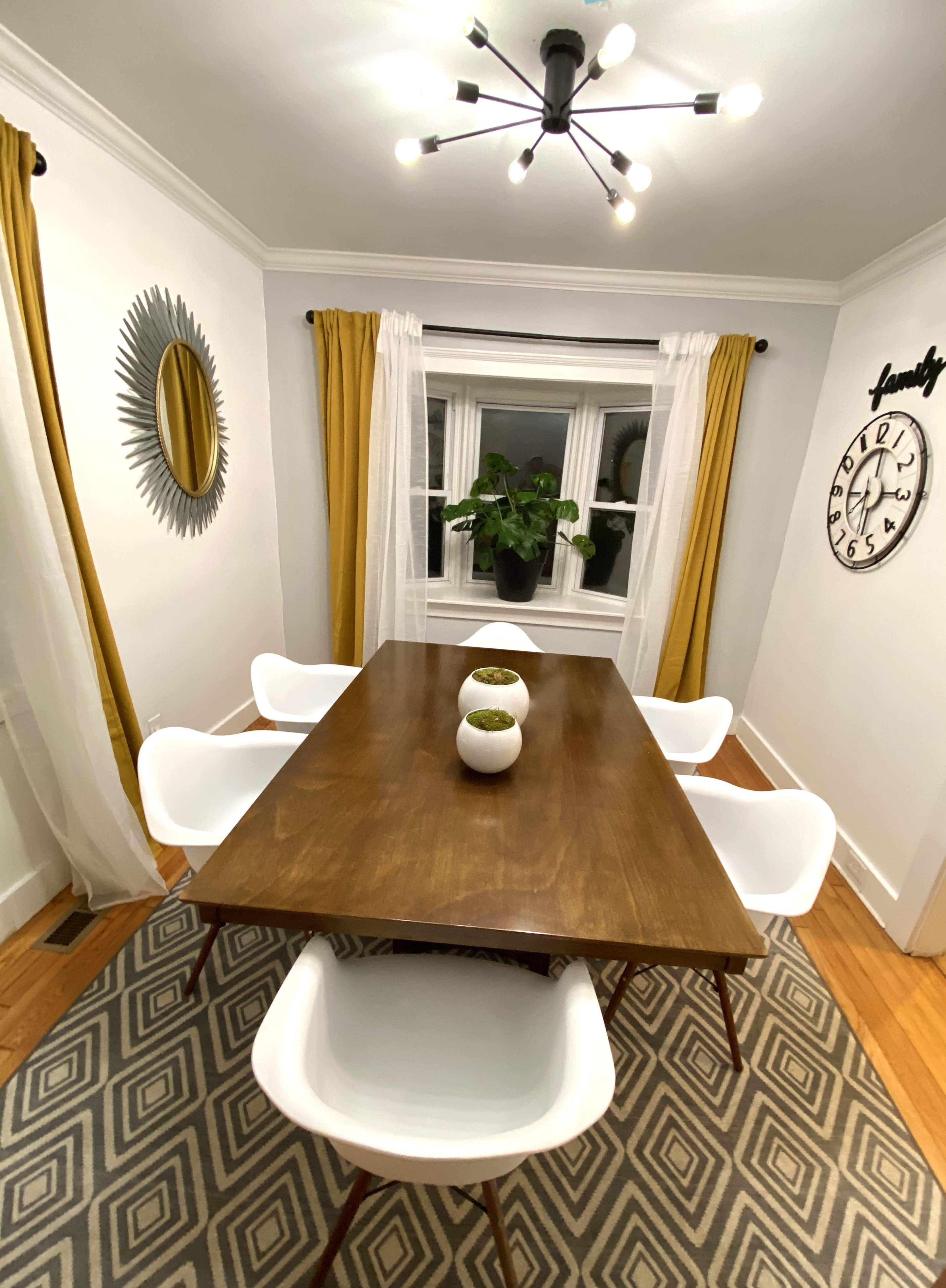 A dining area features a wooden table surrounded by white chairs, with a large window adorned with yellow curtains and a potted plant, alongside a wall clock and a mirror.