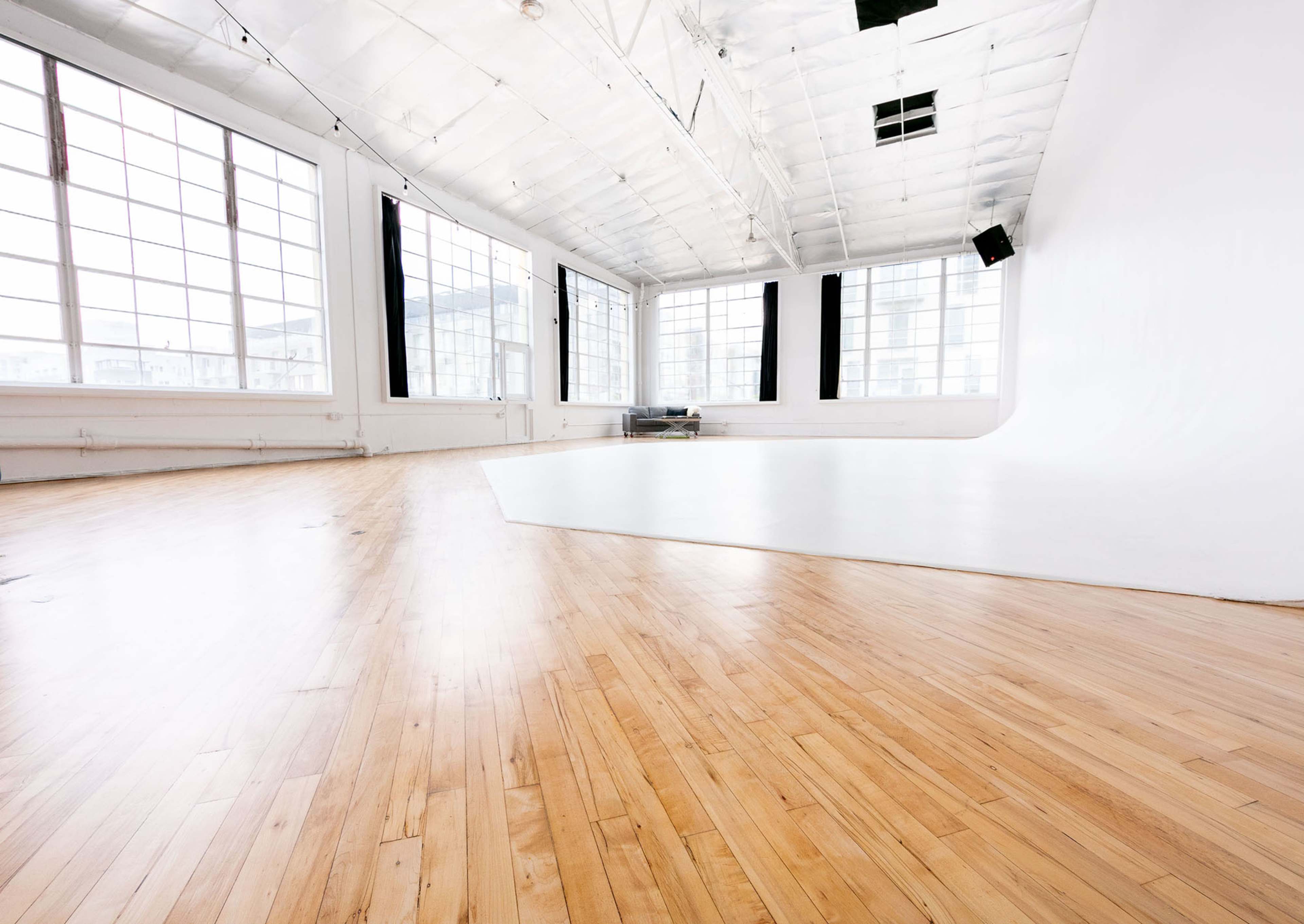 The image shows a spacious studio with wooden flooring and large windows letting in natural light, featuring a white backdrop in one corner.