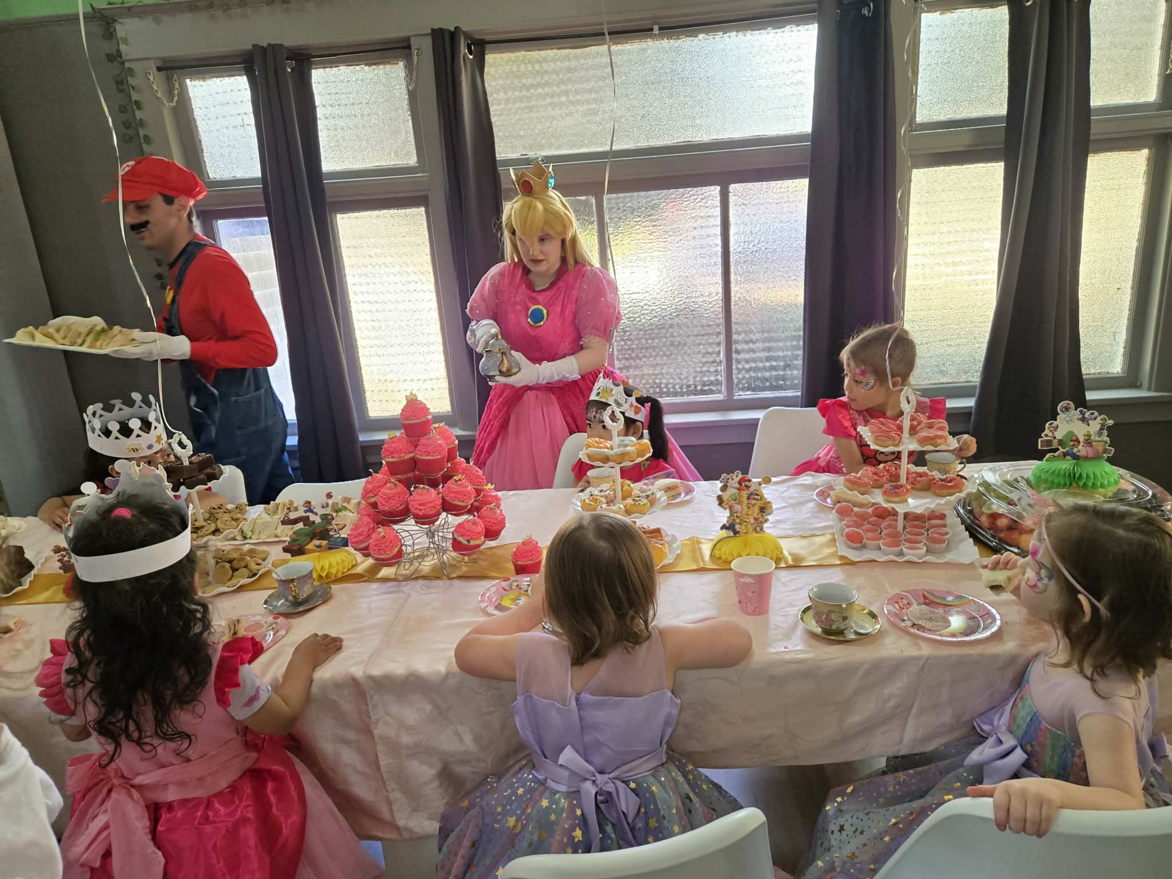 A group of children dressed in princess costumes sits at a table adorned with desserts, while a man in a Mario costume and a woman dressed as Princess Peach interact nearby.