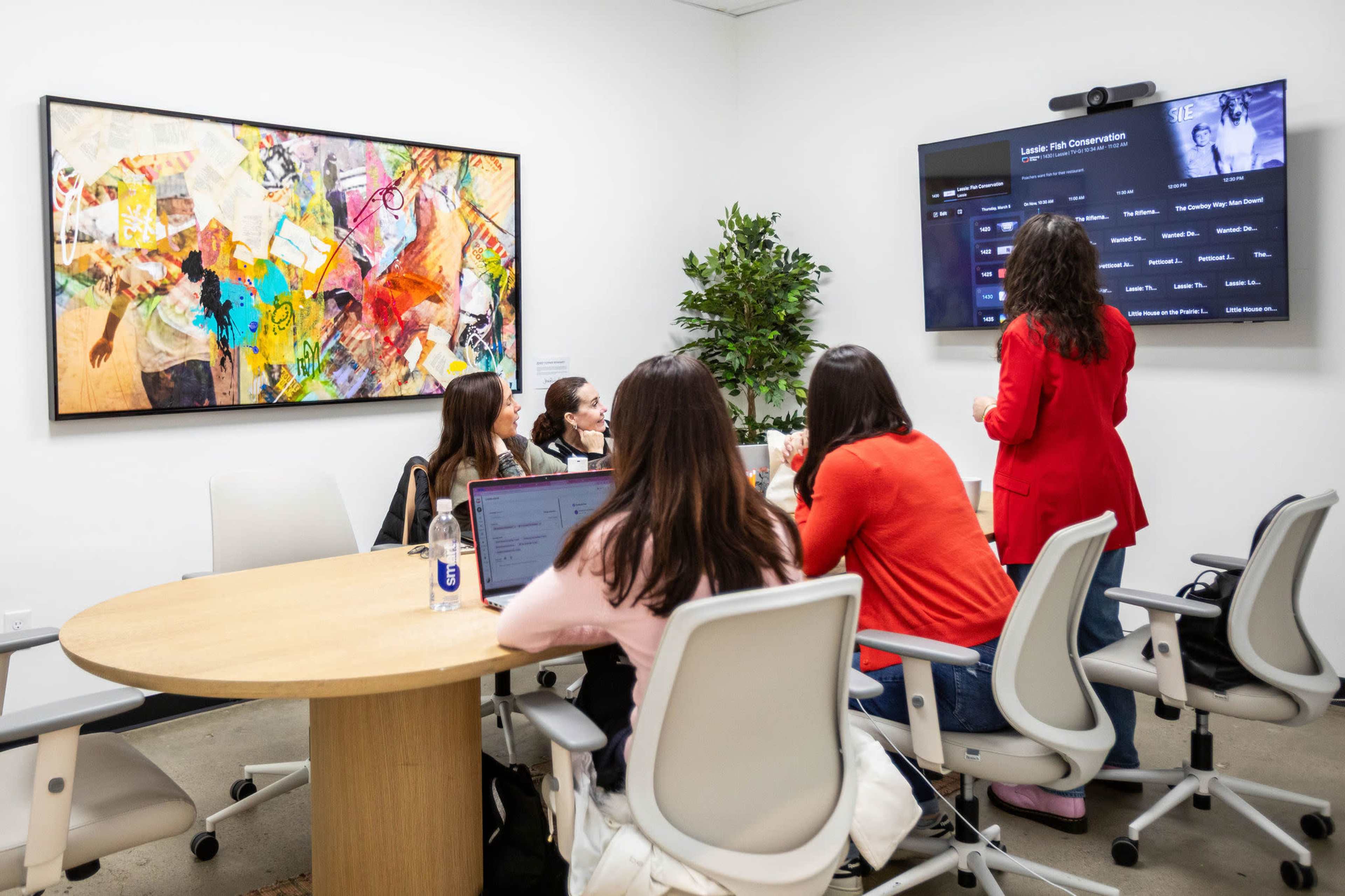 A group of people is seated around a large table in a conference room while one person stands and presents information on a screen.