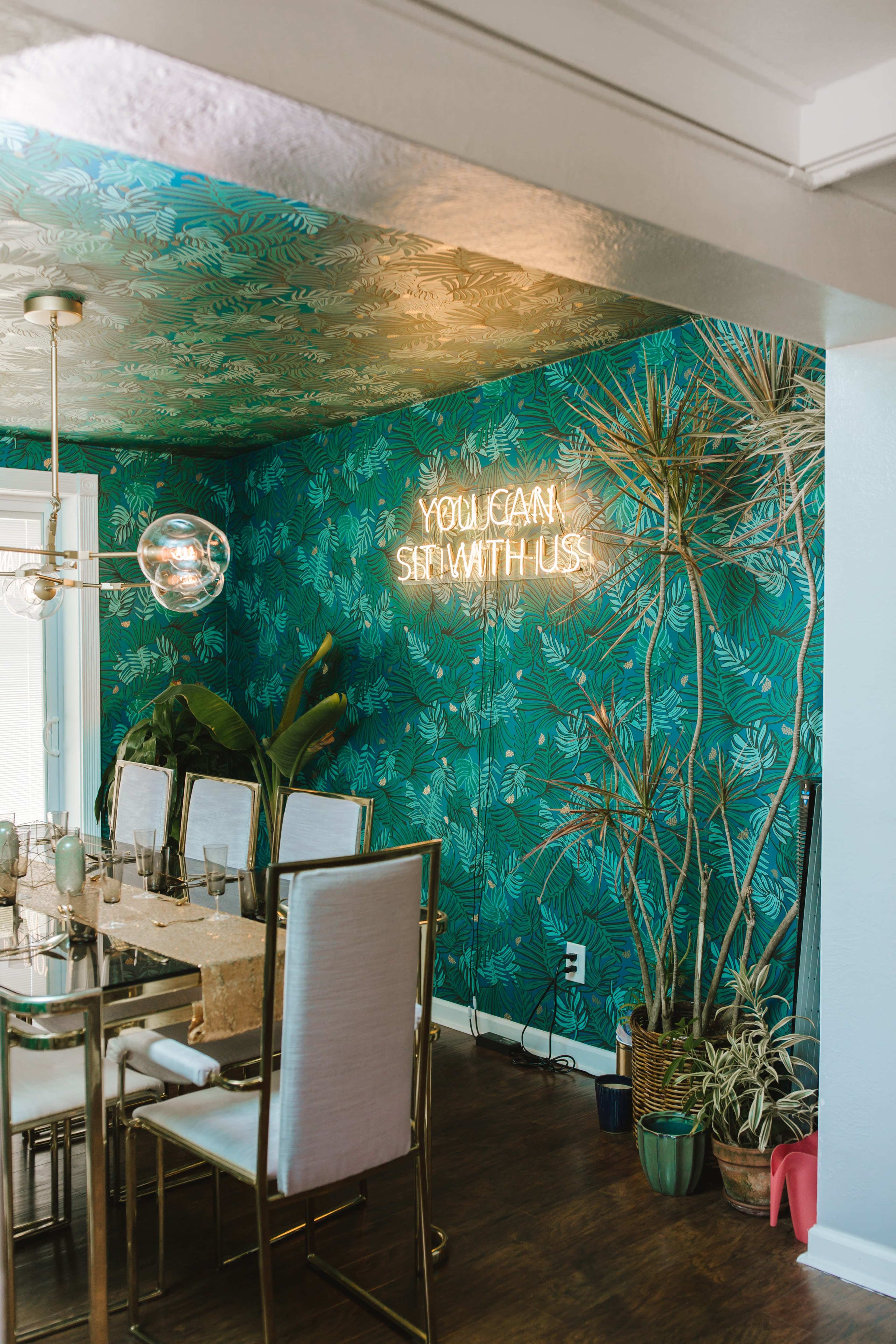 A dining area with a tropical-themed wall, a glass dining table, and a neon sign that reads "YOU CAN SIT WITH US."